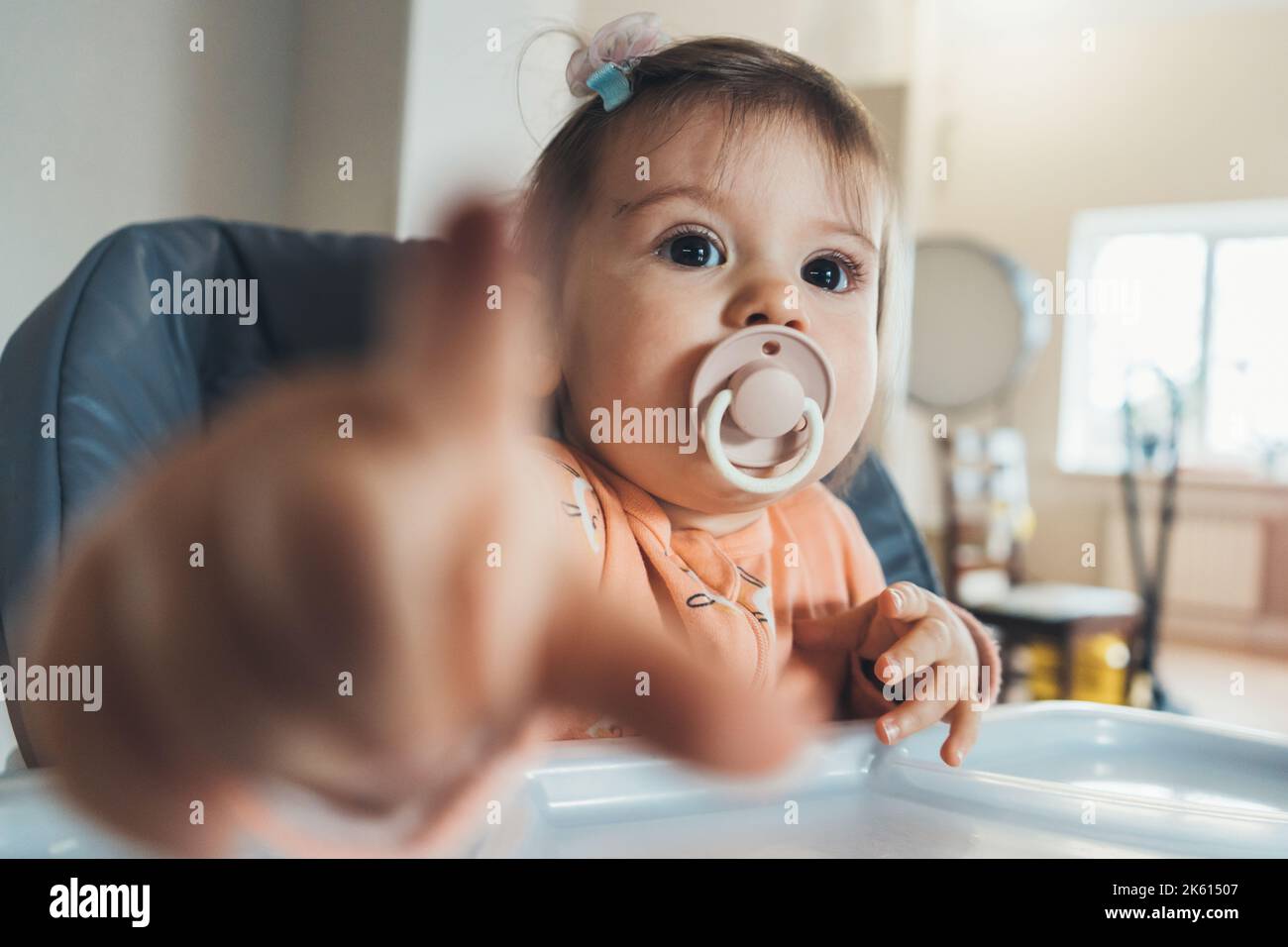 Happy Baby Mädchen auf einem Hochstuhl für die Fütterung von Kindern saugen den Schnuller und zeigt auf die Kamera mit seinem Finger, etwas erkannt Stockfoto