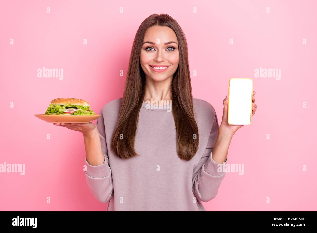 Foto von attraktiven positiven Person Hand halten Burger Platte zeigt leeren Raum Telefon-Bildschirm isoliert auf rosa Hintergrund Stockfoto