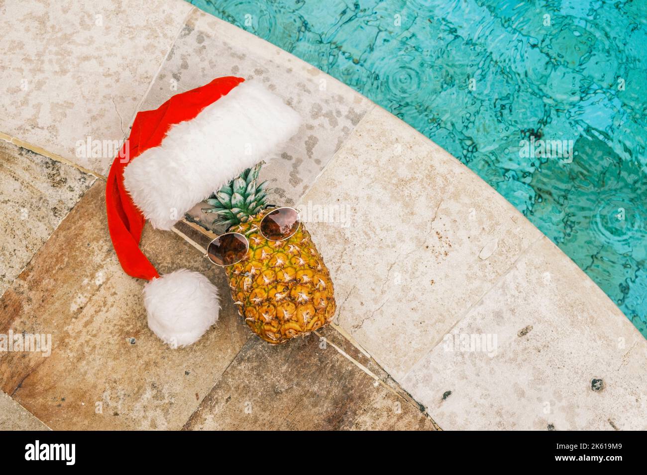 Lustige Ananas mit Sonnenbrille und Weihnachtsmann Hut gegen Pool und tropische Pflanzen bei sonnigem Wetter in den Tropen. Weihnachten in den Tropen. Winter Stockfoto