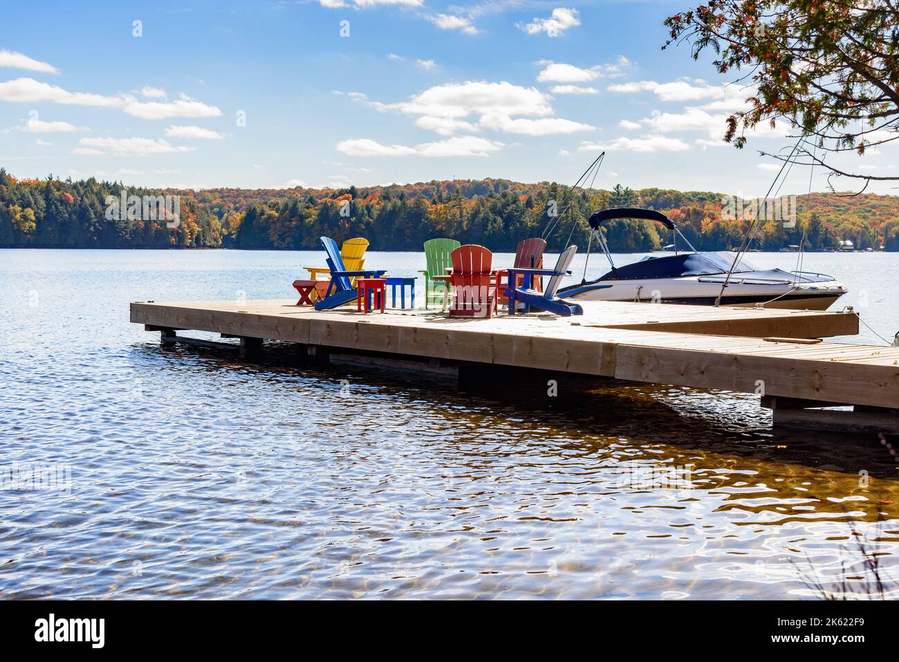Motorboot an einem sonnigen Herbsttag an einem hölzernen Pier am Ufer eines Sees befestigt. Am Pier befinden sich farbenfrohe Sessel. Stockfoto