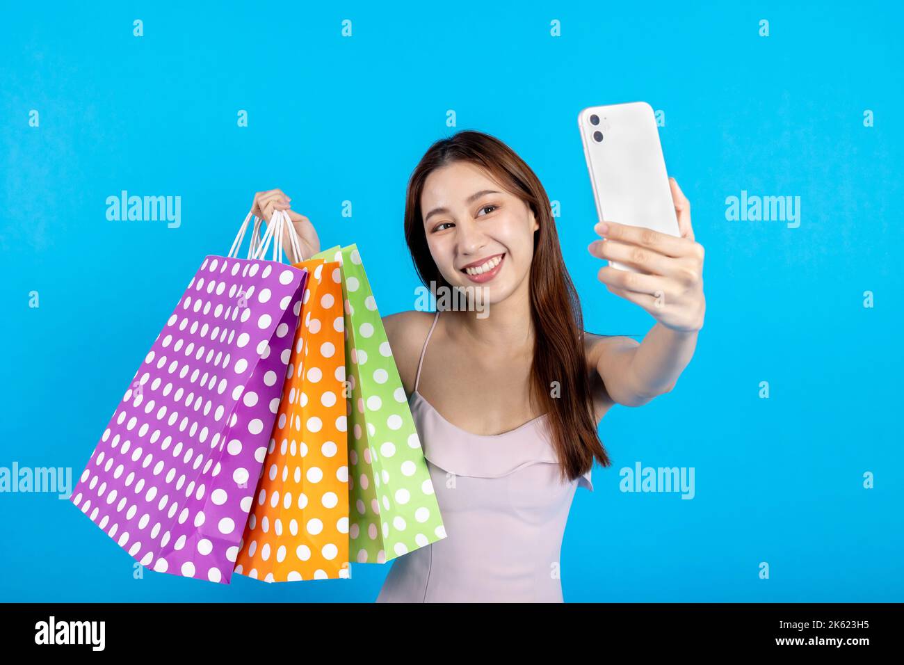 Porträt einer glücklich lächelnden asiatischen Frau im Sommerkleid mit Paketen Einkaufstaschen mit Einkäufen. Selfie nach dem Einkaufen auf Handy isoliert auf blauem Hintergrund - Platz für Werbung kopieren Stockfoto