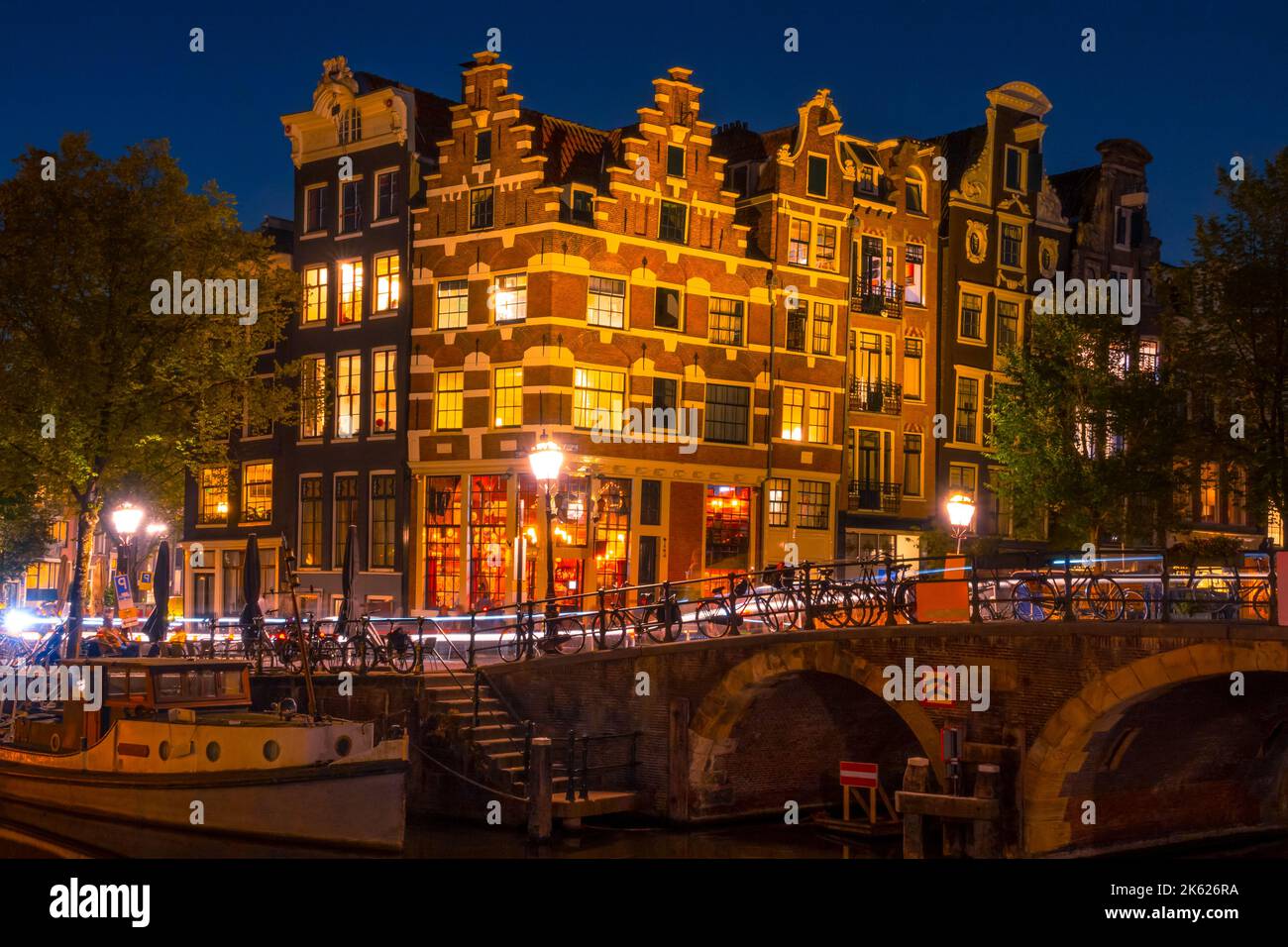 Niederlande. Nacht auf dem Amsterdamer Kanal. Authentisches holländisches Haus mit Lichtern. Leichte Wege von Autos auf der Brücke. Altes Boot, das festgemacht wurde Stockfoto