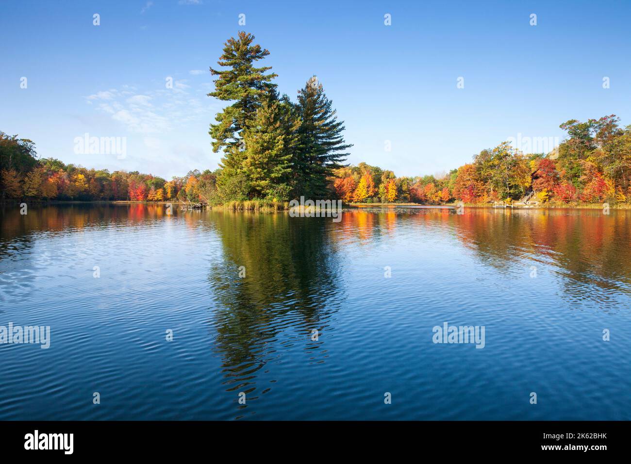 Schöner blauer See mit Bäumen in Herbstfarbe und einer kleinen Insel an einem hellen Morgen Stockfoto
