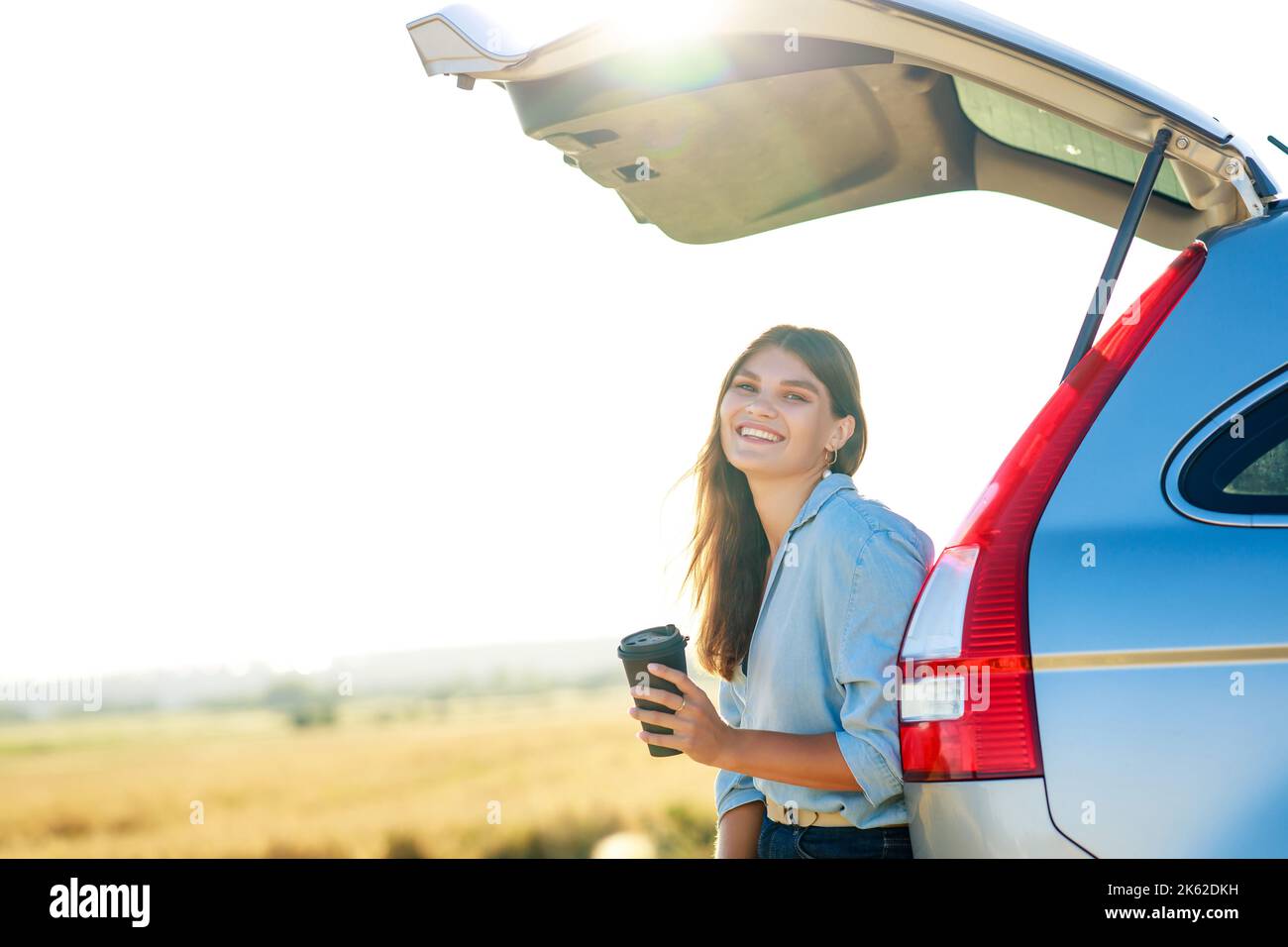 Glückliche junge Frau, die Kaffeetasse hält und bei Sonnenuntergang auf dem offenen Kofferraum ihres Autos in einem Weizenfeld sitzt Stockfoto