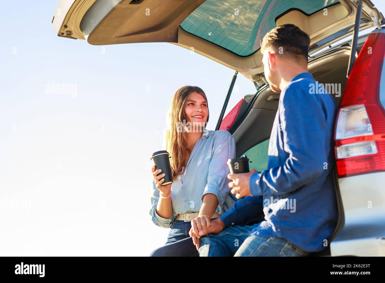 Das junge Paar, der Mann und die Frau, die mit einem neuen Auto unterwegs sind, halten bei Sonnenuntergang auf einem Weizenfeld an, um Kaffee zu trinken Stockfoto