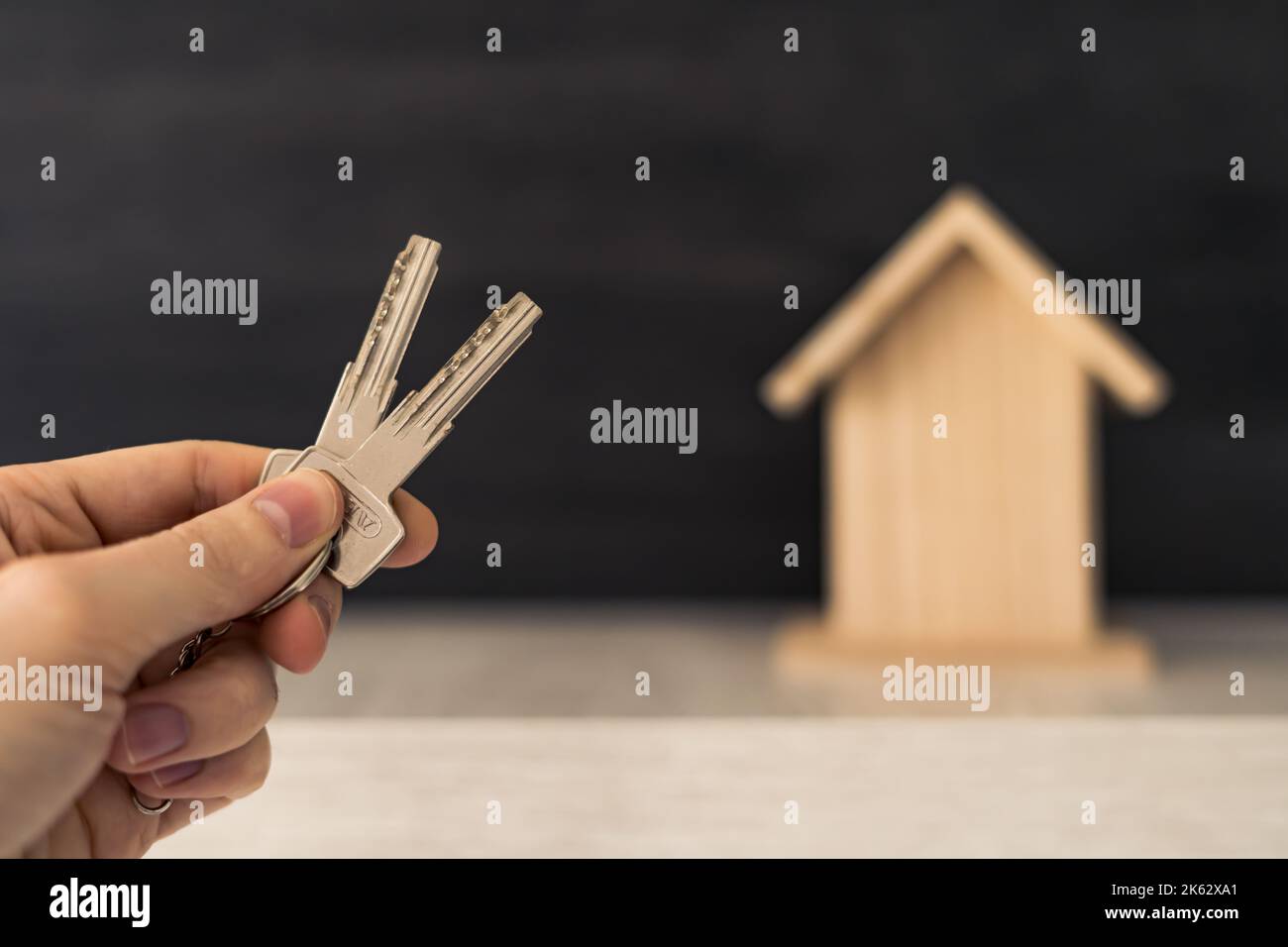 Die Hand einer Person, die die Schlüssel für ihr neues Zuhause hält, mit einem kleinen Holzhaus im Hintergrund. Stockfoto