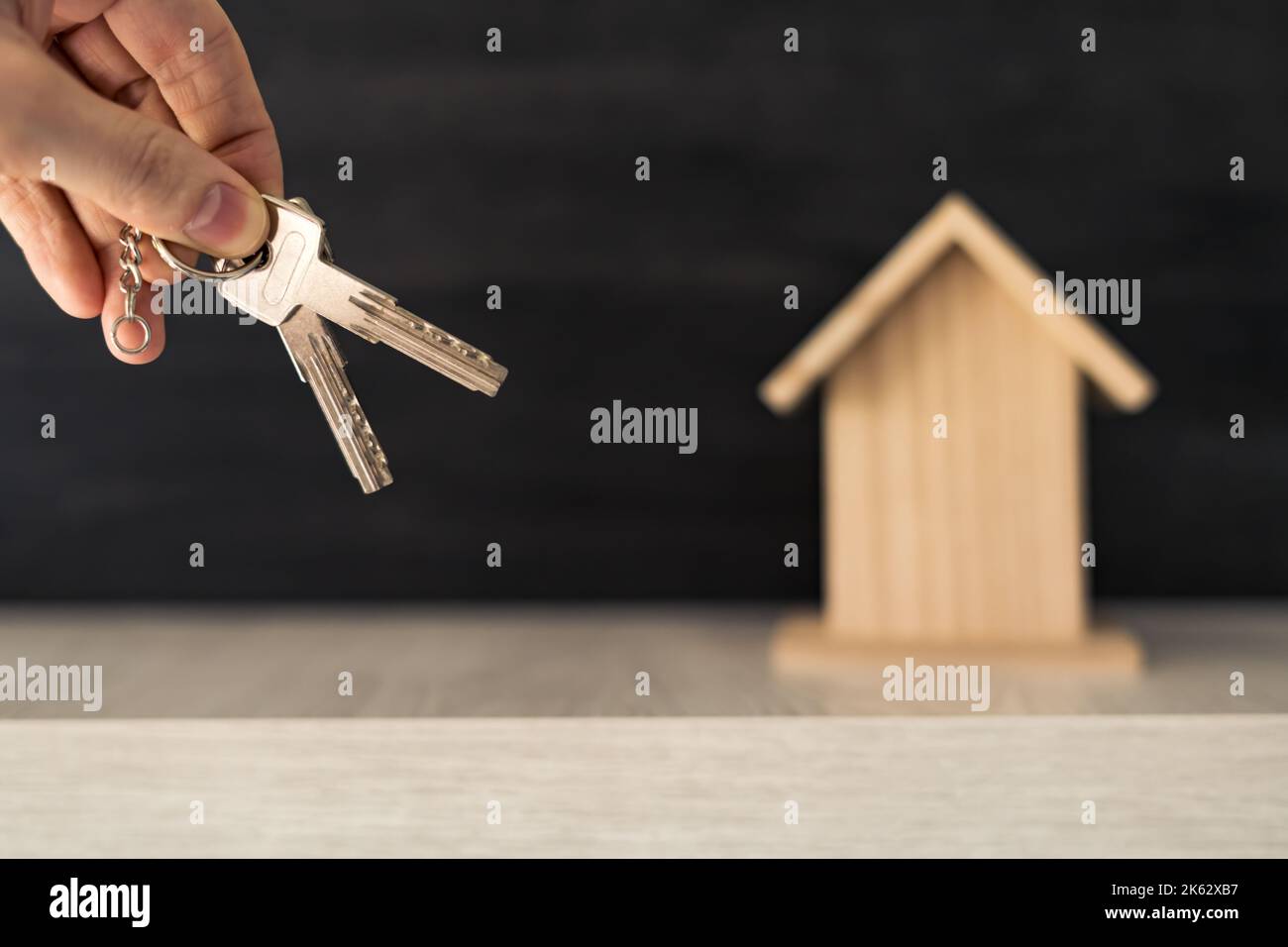 Die Hand einer Person, die die Schlüssel für ihr neues Zuhause hält, mit einem kleinen Holzhaus im Hintergrund. Stockfoto