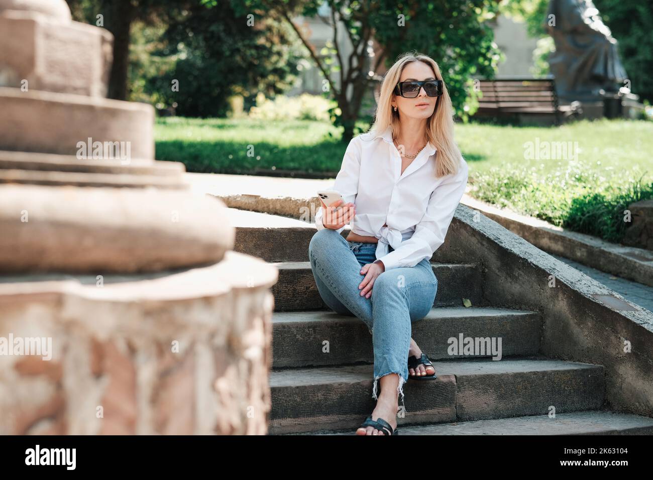 Elegante kaukasische Frau, die auf der Treppe im öffentlichen Park sitzt und Smartphone in der Hand hält, selbstbewusste Unternehmerin, die eine Sonnenbrille trägt Stockfoto
