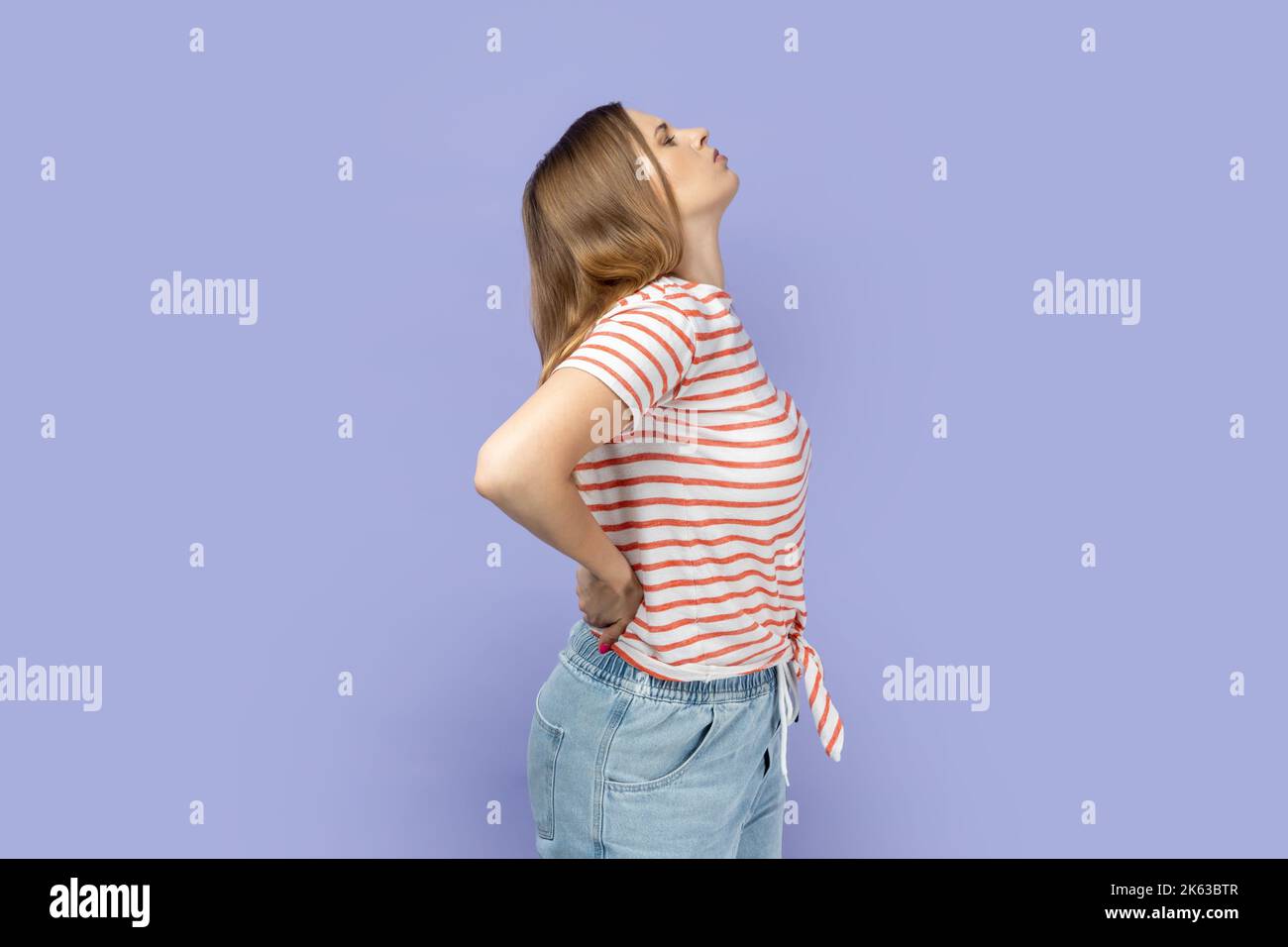 Seitenansicht einer blonden Frau, die gestreiftes T-Shirt trägt, das den Rücken berührt und vor Schmerzen schreit, Rückenschmerzen leidet, Beschwerden an der unteren Lendenwirbelsäule hat. Innenaufnahme des Studios isoliert auf violettem Hintergrund. Stockfoto