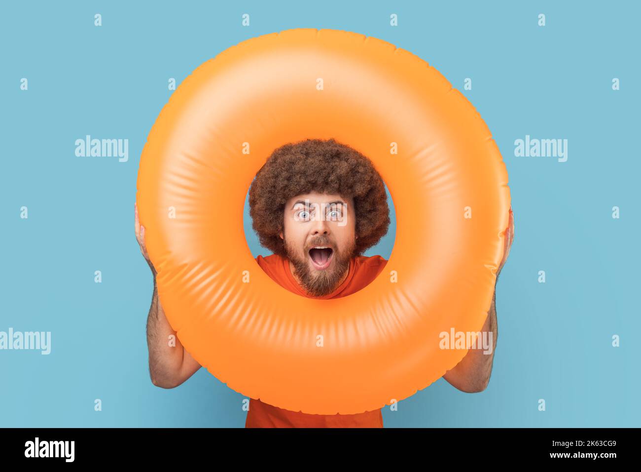 Porträt eines Mannes mit schockierter Gesichtsausdruck, der durch einen leuchtend orangefarbenen Gummiring schaut, die Kamera mit offenem Mund anschaut, überrascht vom Urlaub. Innenaufnahme des Studios isoliert auf blauem Hintergrund. Stockfoto