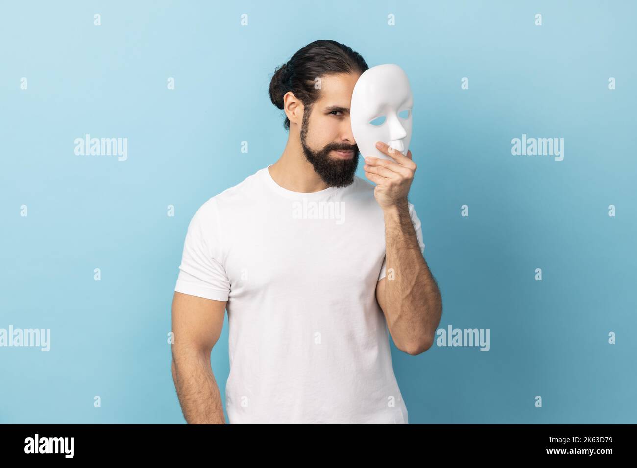 Portrait eines Bodybuilders mit Bart, das weißes T-Shirt trägt und weiße Maske hält, guckt, die Kamera anschaut und vorgibt, eine andere Person zu sein. Innenaufnahme des Studios isoliert auf blauem Hintergrund. Stockfoto