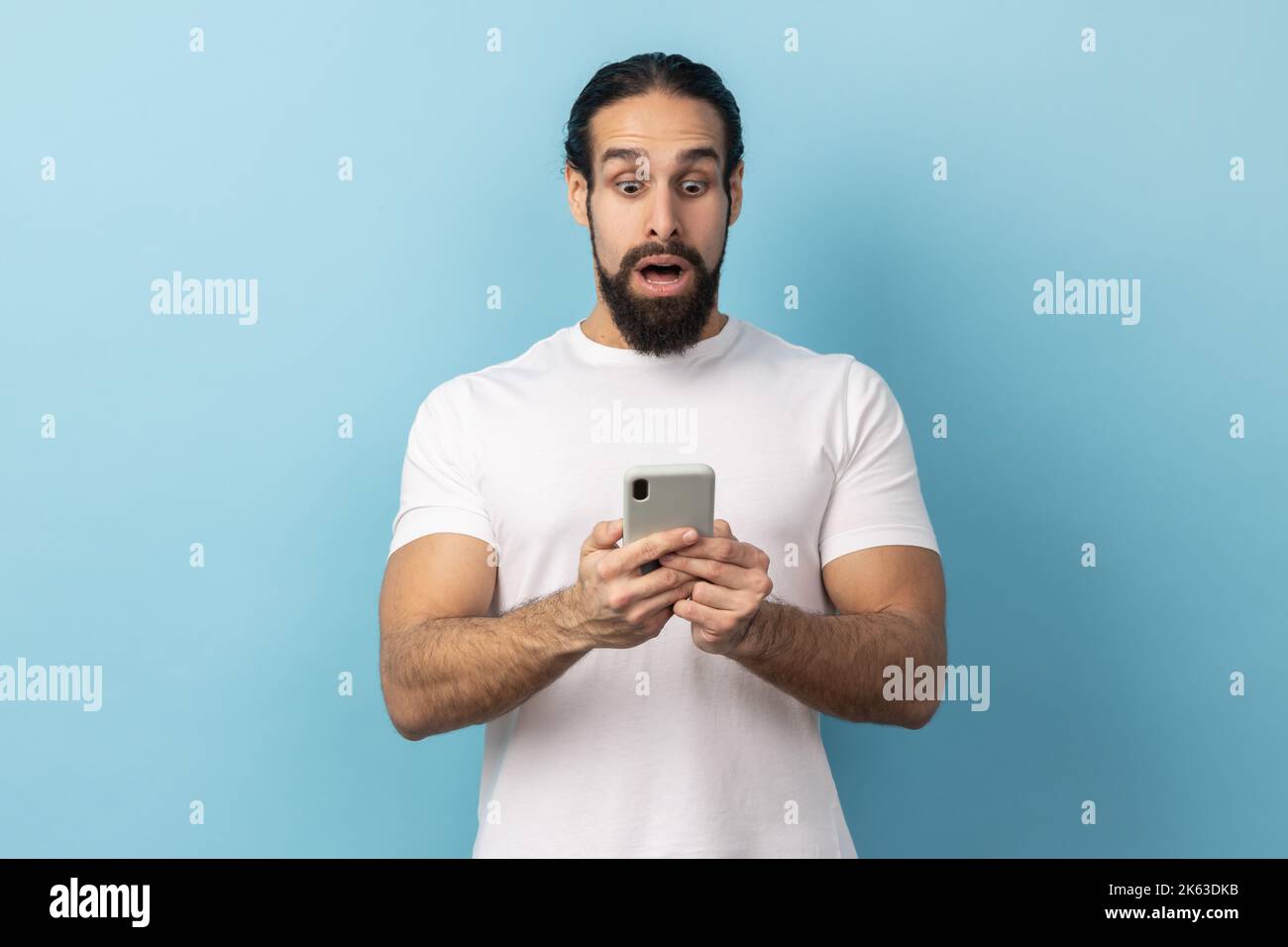 Porträt eines überraschten Mannes mit Bart und weißem T-Shirt, der einen Schock ausdrückt, während er das Handy benutzt, im Internet sucht und erstaunliche Nachrichten zeigt. Innenaufnahme des Studios isoliert auf blauem Hintergrund. Stockfoto