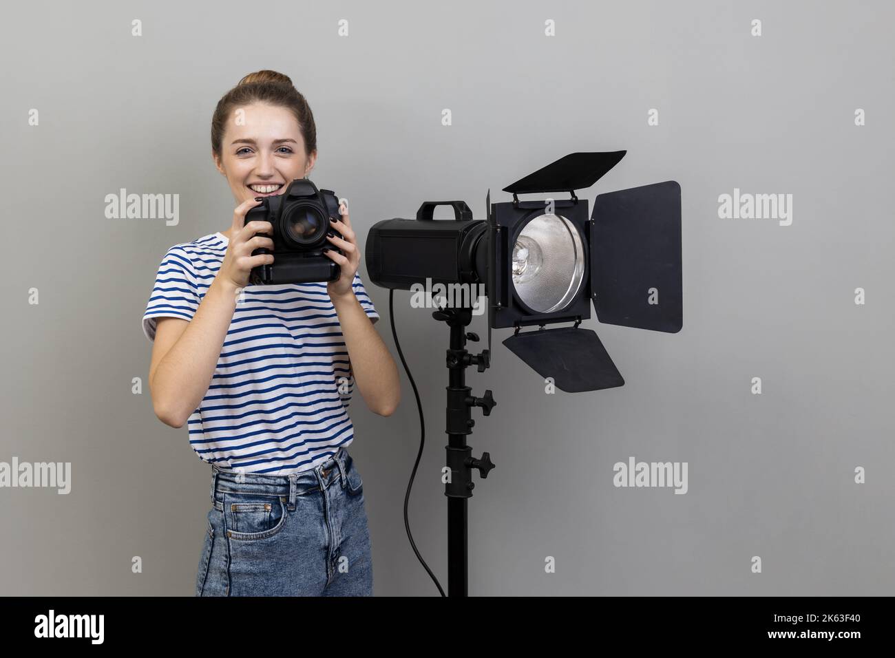 Porträt einer lächelnden, zufriedenen Fotografin mit gestreiftem T-Shirt, die mit einer Fotokamera und einem Reflektor steht und die Kamera anschaut. Innenaufnahme des Studios isoliert auf grauem Hintergrund. Stockfoto