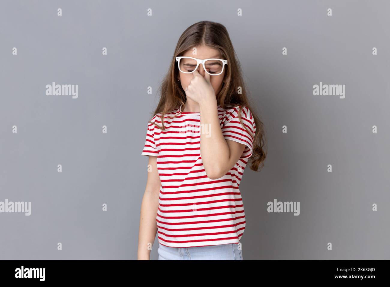 Porträt eines kranken, müden kleinen Mädchens mit gestreiftem T-Shirt, das mit Rippen in den Augen steht, die Brille auszieht, die Augen verletzt und Kopfschmerzen verursacht. Innenaufnahme des Studios isoliert auf grauem Hintergrund. Stockfoto