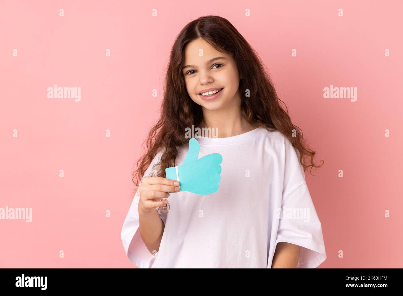 Portrait von lächelnd entzückend entzückende kleine Mädchen trägt weiße T-Shirt halten und zeigt wie oder Daumen nach oben Papier Form Zeichen. Innenaufnahme des Studios isoliert auf rosa Hintergrund. Stockfoto