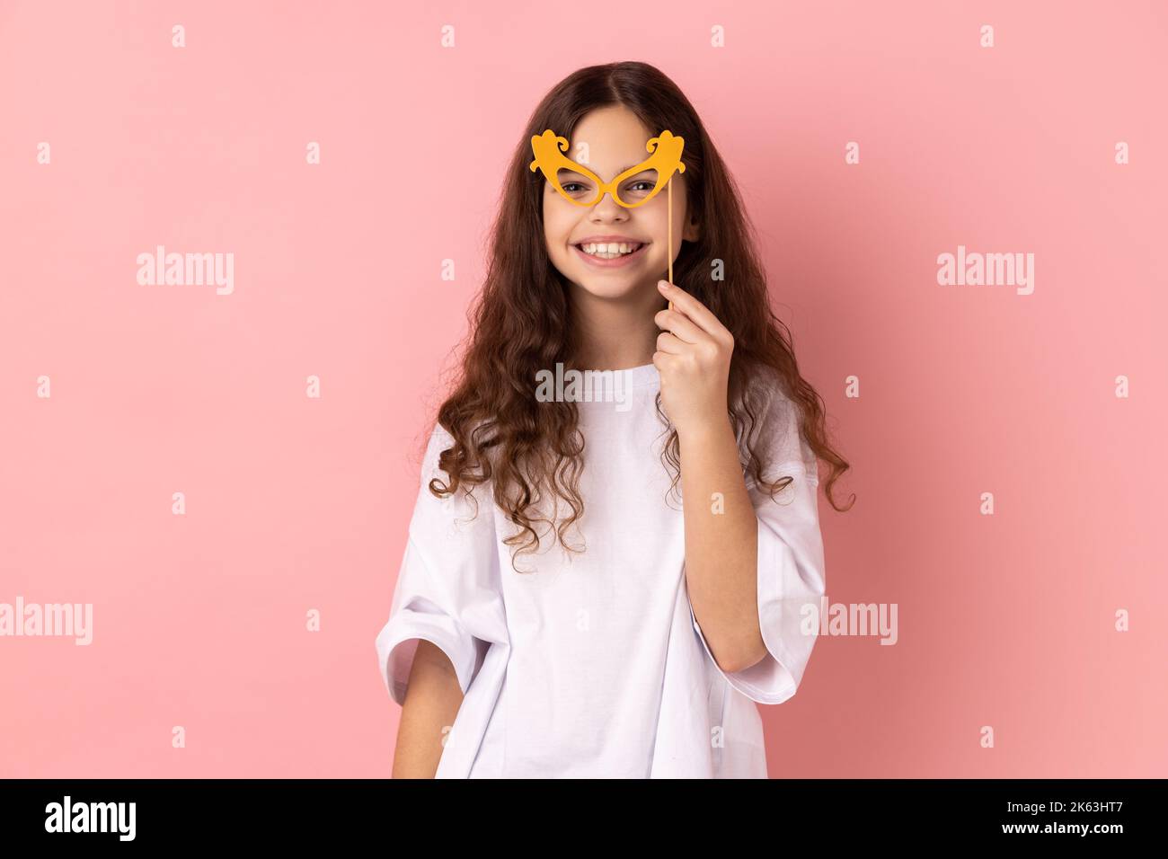 Portrait of cute happy Little girl tragend white T-Shirt lächelnd und halten Papiergläser auf Stick, mit Party-Requisiten im Urlaub, Spaß haben. Innenaufnahme des Studios isoliert auf rosa Hintergrund. Stockfoto