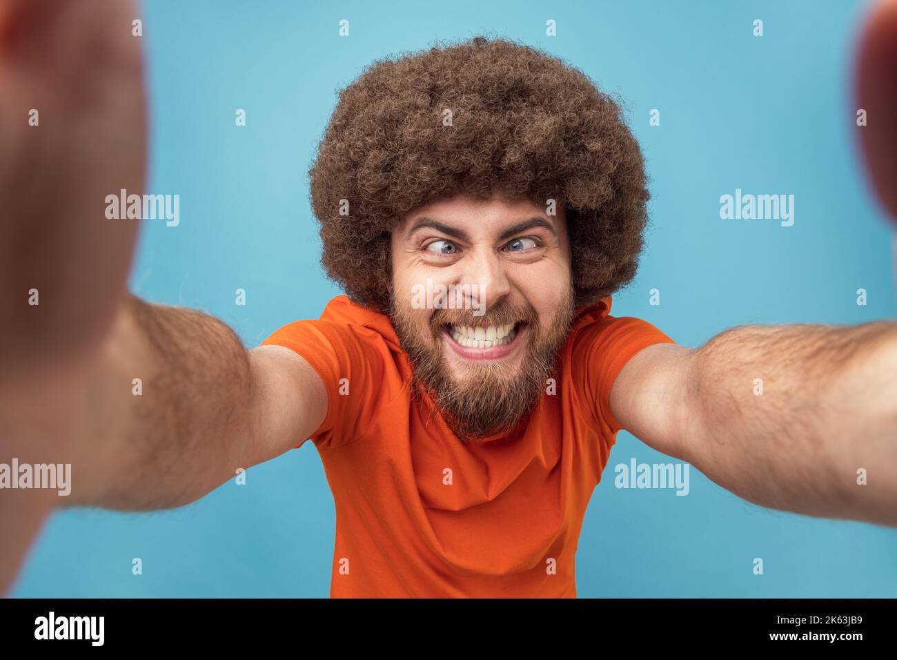 Porträt eines verrückten albernen Mannes mit Afro-Frisur in orangefarbenem T-Shirt, der Livestream überträgt, Spaß mit Abonnenten hat, mit gekreuzten Augen posiert, POV. Innenaufnahme des Studios isoliert auf blauem Hintergrund. Stockfoto