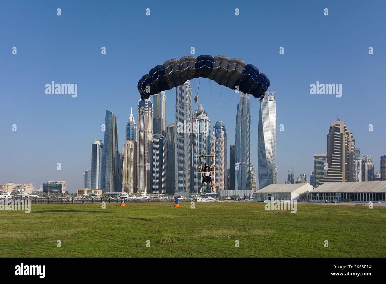 Dubai, Vereinigte Arabische Emirate - Tandem-Fallschirmspringer bereiten sich darauf vor, mit einem Fallschirm zurück in die Palm Drop Zone zu landen. Dubai Marina Wolkenkratzer im Hintergrund. Stockfoto