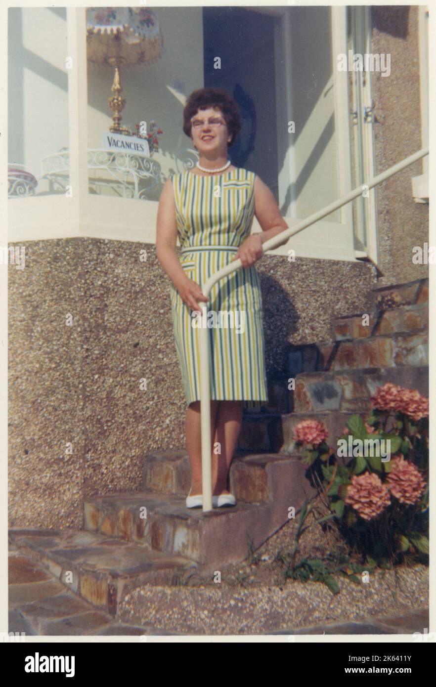 Frau mittleren Alters, die auf einer Reihe von Stufen steht, die bis zur Haustür eines Hauses führen, möglicherweise am Meer - September 1963 Stockfoto