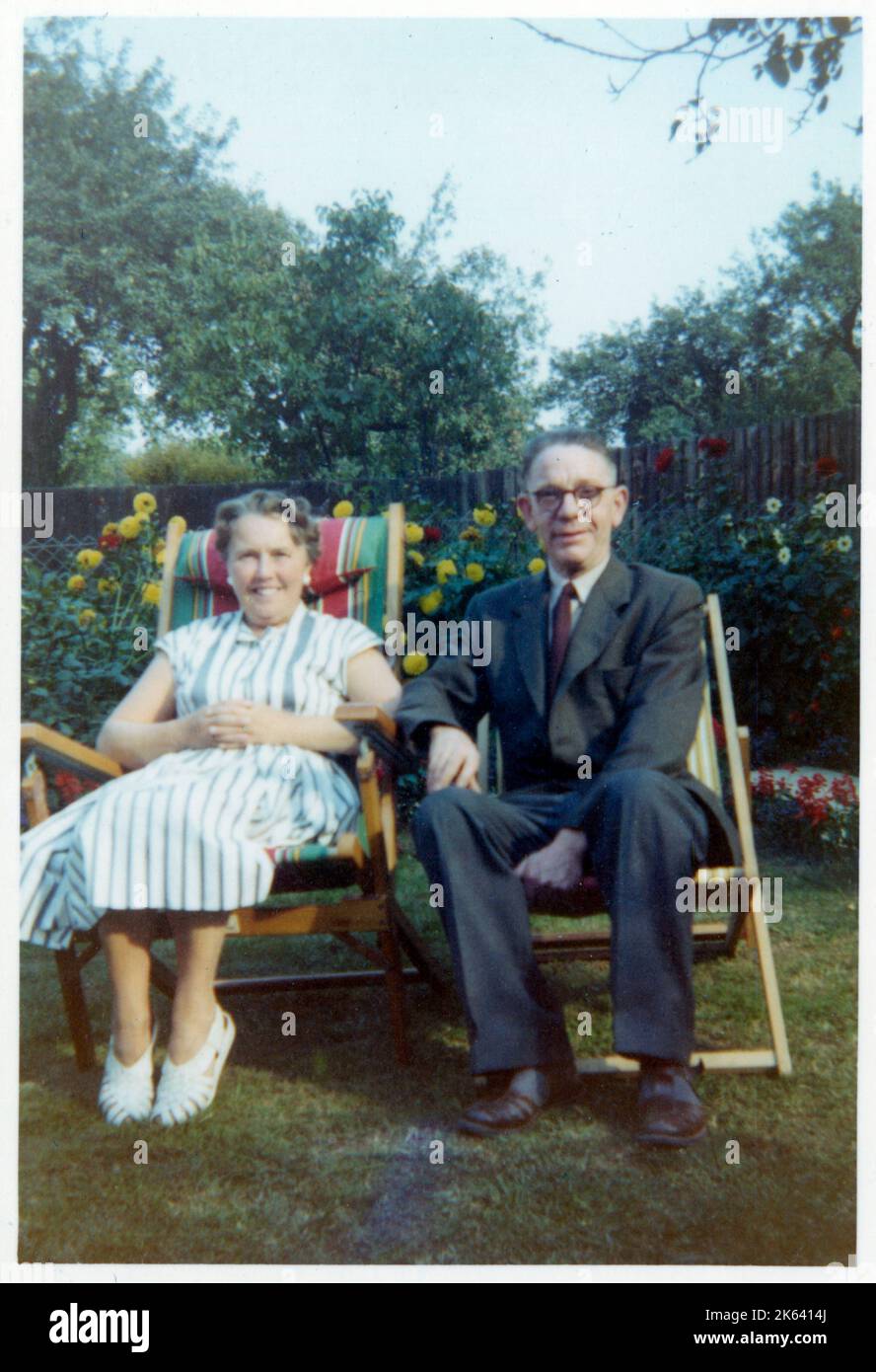 Ein fröhliches älteres Paar, das in leichten ausklappbaren Gartenstühlen aus Holz und Leinwand in einem gepflegten Vorstadtgarten sitzt - September 1959 Stockfoto