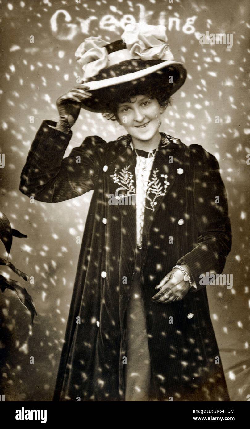 Eine junge Dame in einem großen Hut mit Schleifenbefall und einem eleganten Samtmantel lächelt durch den herabfallenden Winterschnee. Stockfoto