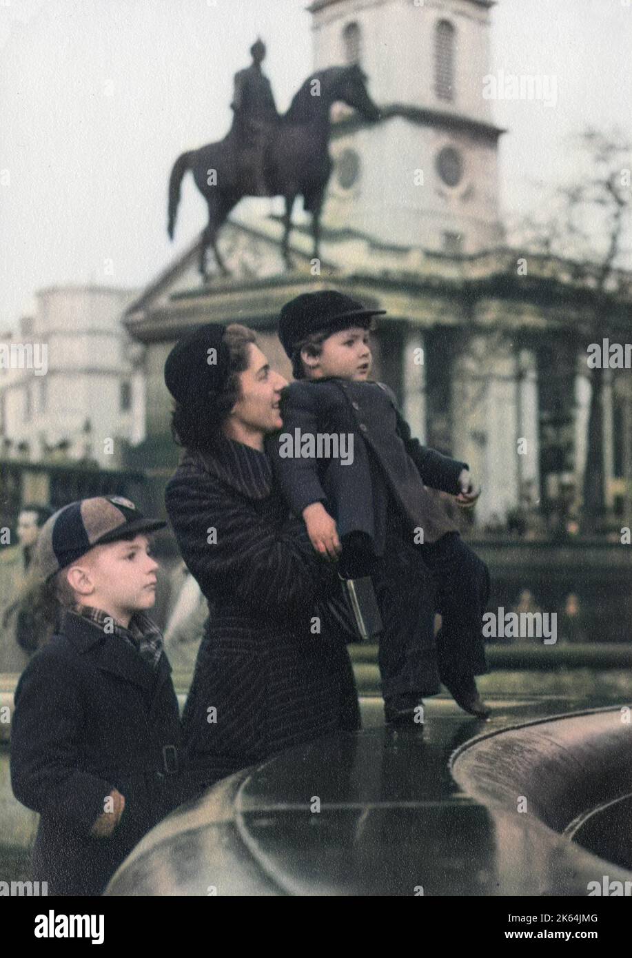 Ein wunderbares Foto von einer Mutter und ihren beiden Jungen bei einem der Brunnen am Trafalgar Square, London - 1950er. Die unverwechselbare Form der Kirche St. Martin-in-the-Fields ist im Hintergrund zu sehen, hinter der Statue von König George IV. Auf dem Pferderücken. Stockfoto