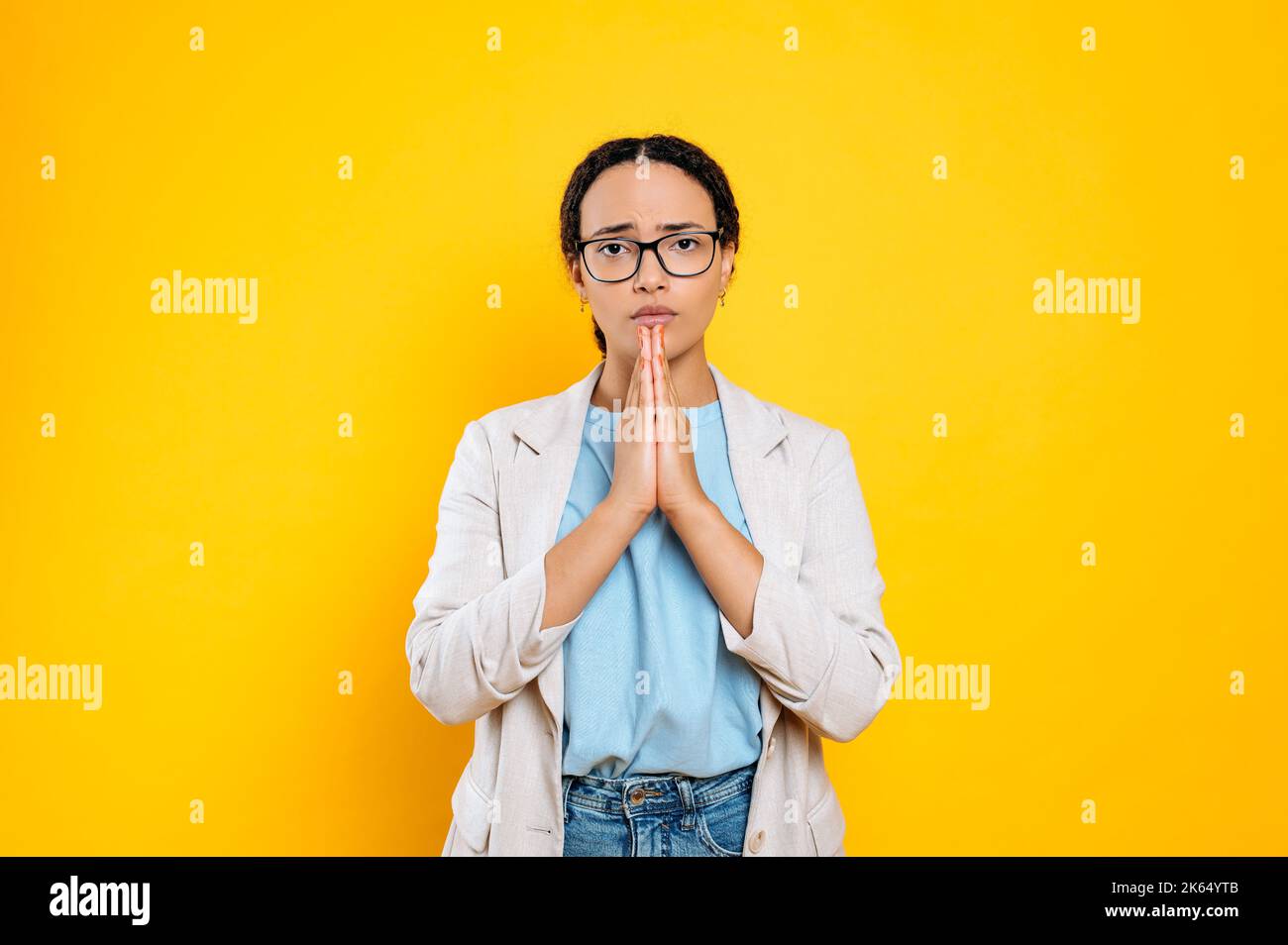 Nach etwas fragen. Flehend süße brasilianische oder latino junge Brünette Frau, Firmenarbeiterin, hält die Hände in einem gefalteten Gebetsschild, schaut auf die Kamera, steht auf isoliertem orangefarbenem Hintergrund Stockfoto