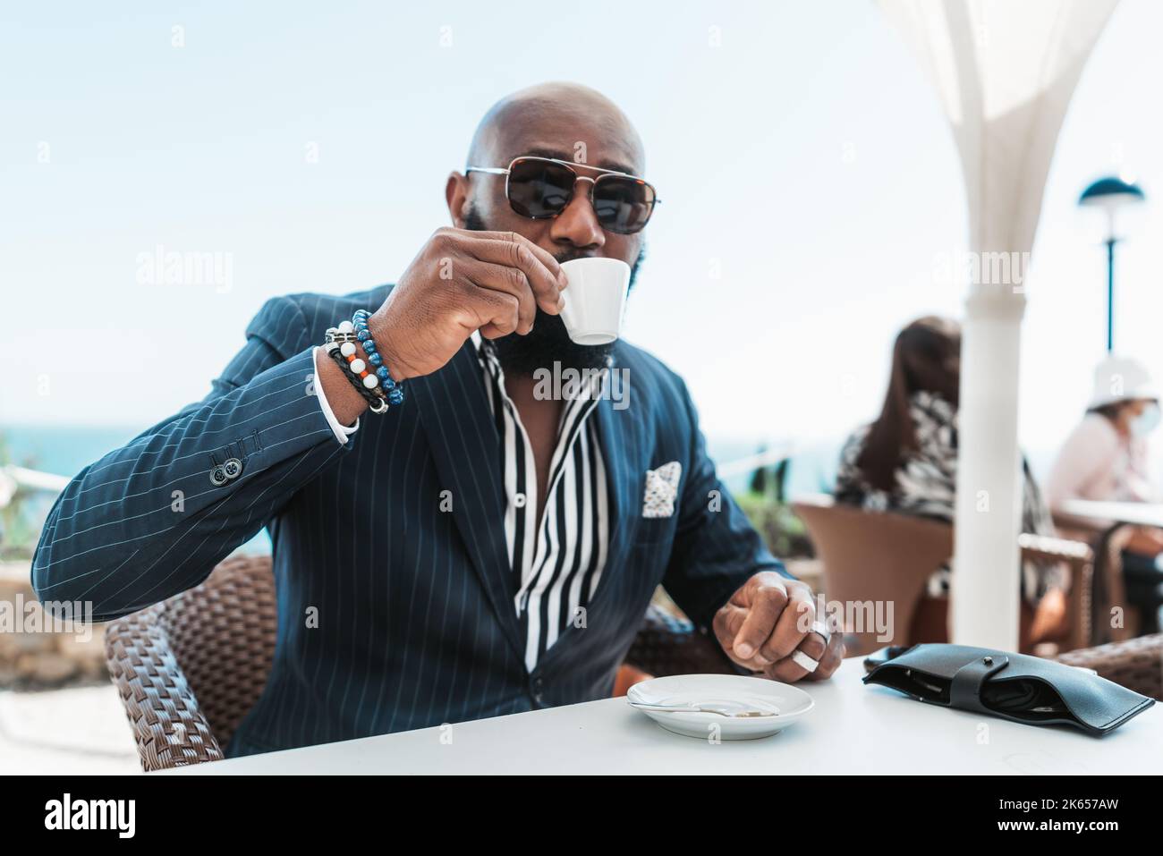 Ein Porträt eines kahlen Afrikaners in Sonnenbrille und eines modischen Sommerkanzers, der in einem Straßencafé sitzt und Kaffee trinkt, mit einem selektiven FOC Stockfoto