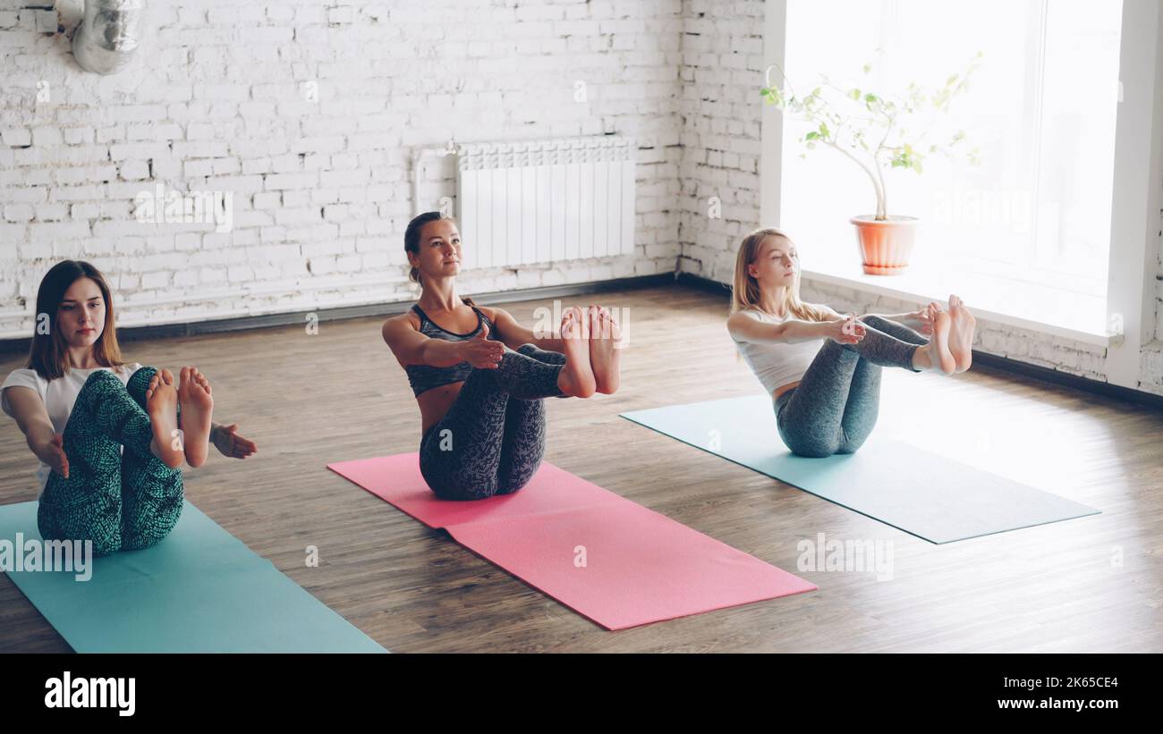 Pan Shot von schlanken jungen Damen, die Yoga üben und dabei eine Balancierbootpose in einem hellen, geräumigen Raum machen. Junge Frauen konzentrieren sich auf Gegengewicht, sie üben gerne. Stockfoto