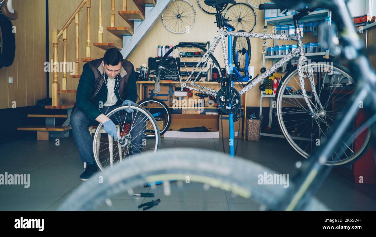 Hübscher männlicher Mechaniker fixiert das Hinterrad mit einem Schraubenschlüssel, während er am modernen Arbeitsplatz auf einem Holzhocker sitzt. Zyklen, Ersatzteile, Werkzeuge und Geräte sind sichtbar. Stockfoto