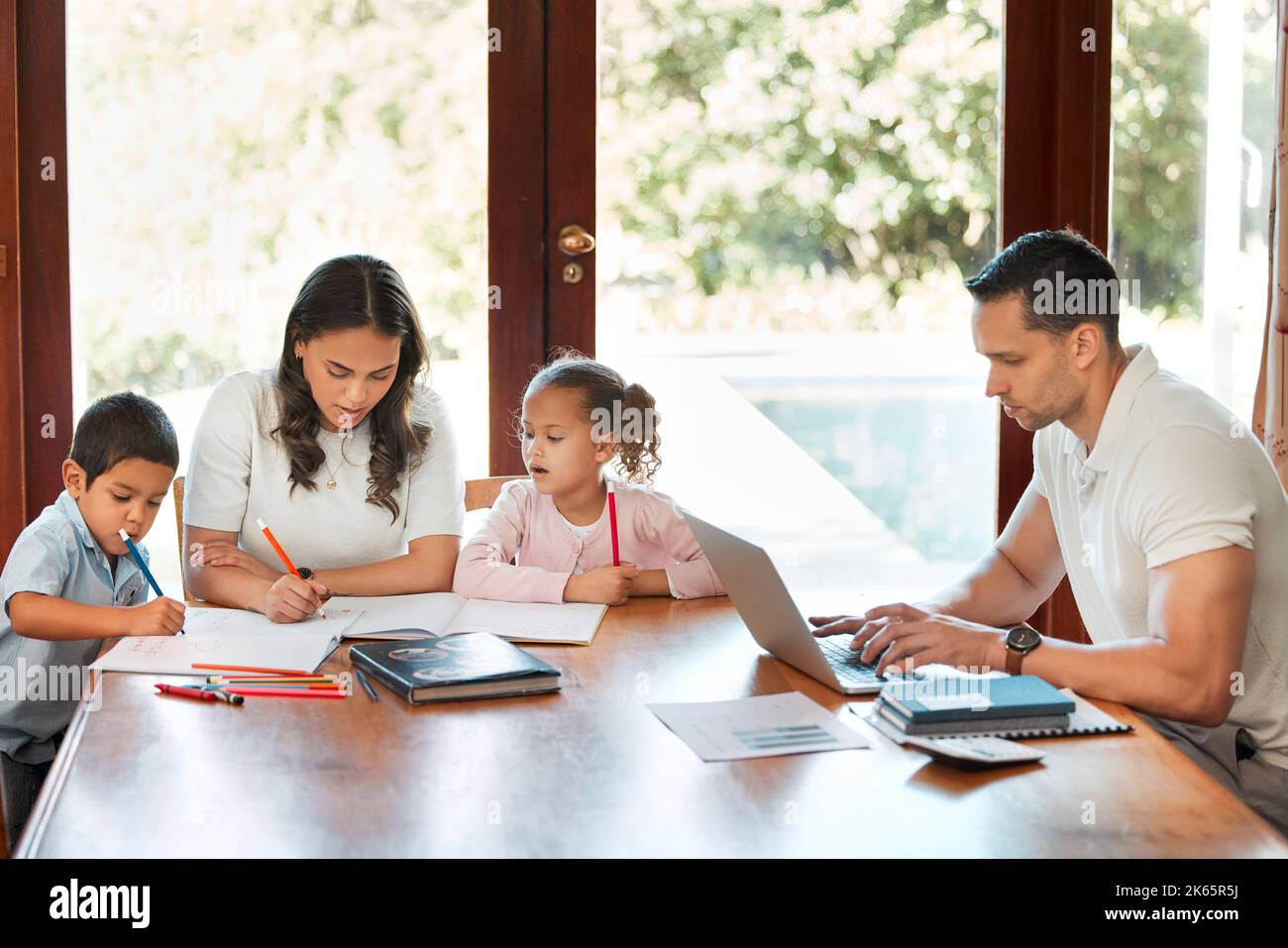 Die junge Mutter aus gemischter Rasse hilft ihren Kindern bei den Hausaufgaben, während ihr Vater an einem Tisch in der Lounge auf einem Laptop arbeitet. Kleine Geschwister zeichnen Stockfoto