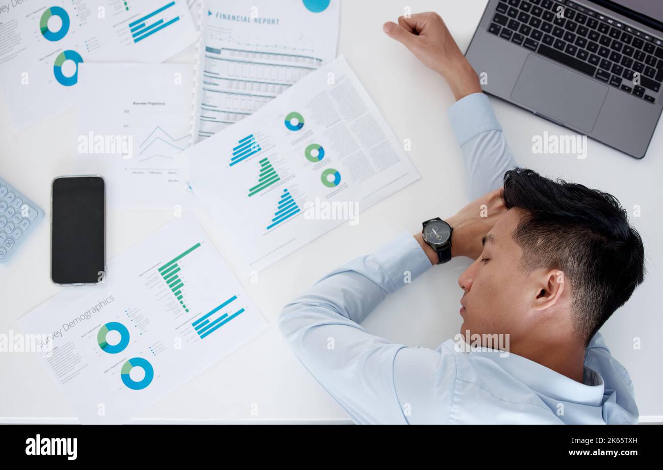 Nahaufnahme eines jungen, müden asiatischen Geschäftsmanns, der auf einem Tisch mit Papierkram und drahtloser Technologie schlief. Junger Mann, der mit einem Laptop erschöpft aussieht Stockfoto