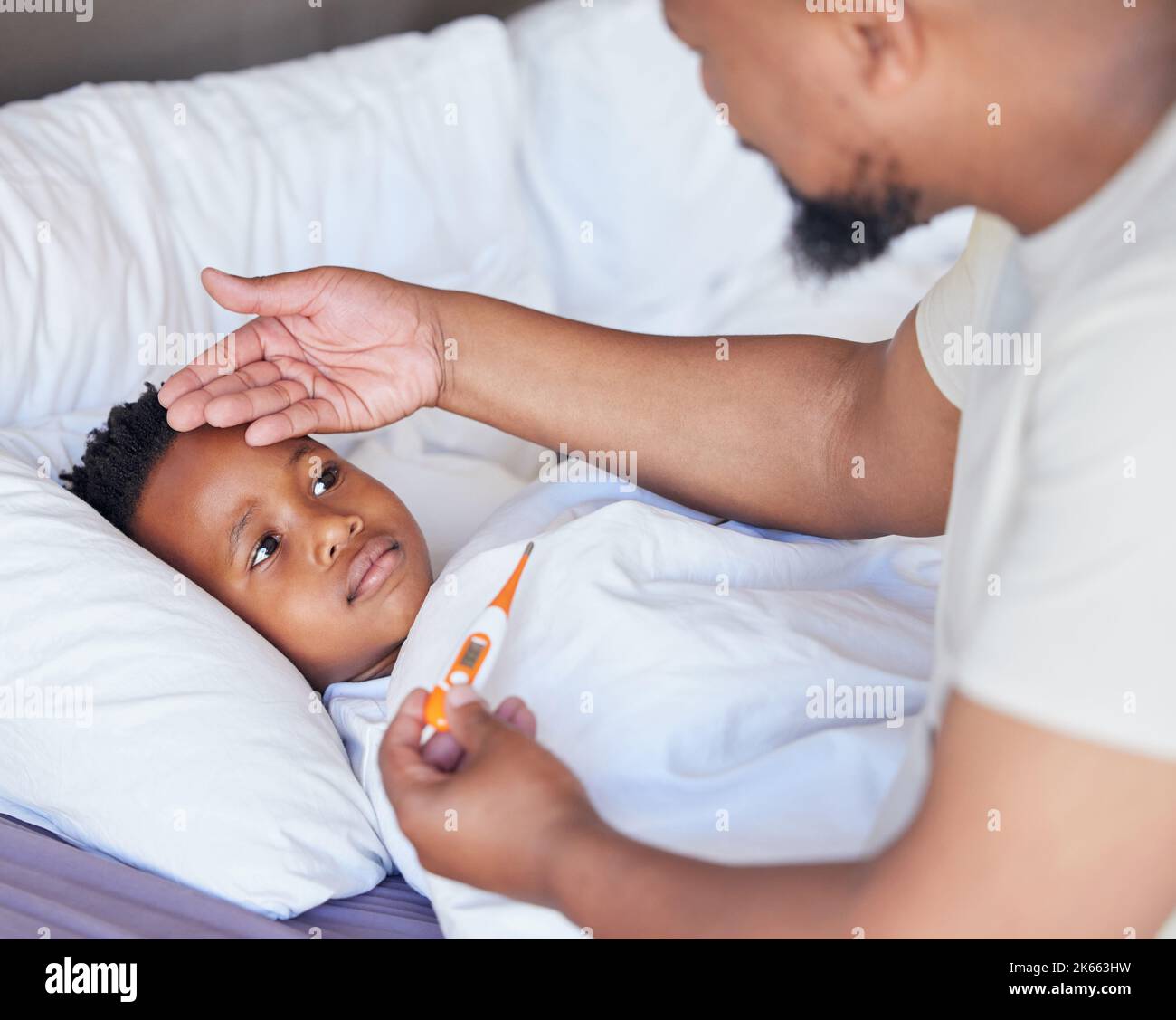 Kranker kleiner Junge im Bett, während sein Vater mit einem Thermometer seine Temperatur überprüft. Schwarze alleinerziehende Mutter, die Söhne auf der Stirn fühlt. Afroamerikanisches Kind Stockfoto