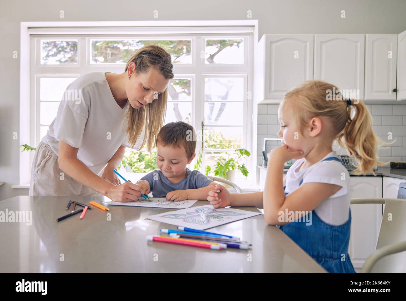Kaukasische Mutter hilft ihren kleinen Kindern, ein Buch mit Farbstiften in der Küche zu Hause zu zeichnen Stockfoto