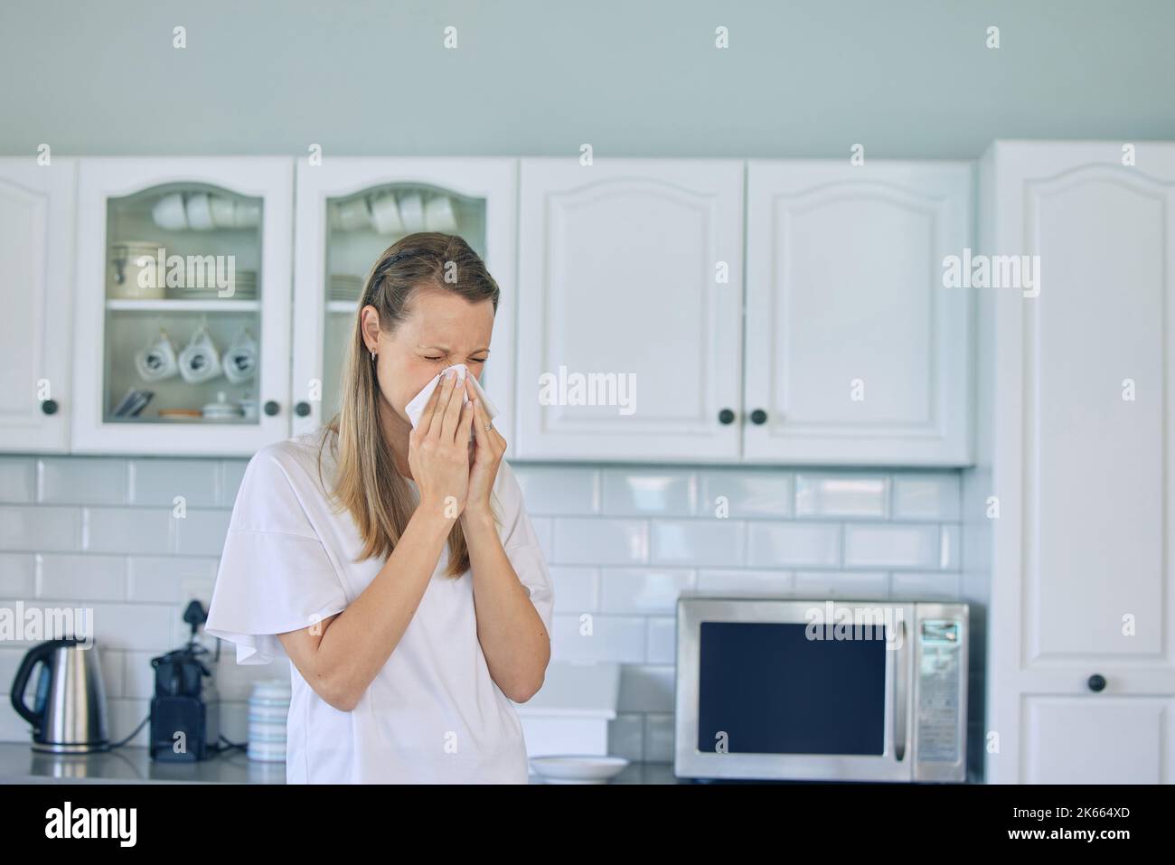 Kranke kaukasische Frau, die zu Hause in der Küche steht und Gewebe hält und ihre Nase bläst. Frau, die an einer saisonalen Allergie oder Grippe leidet Stockfoto