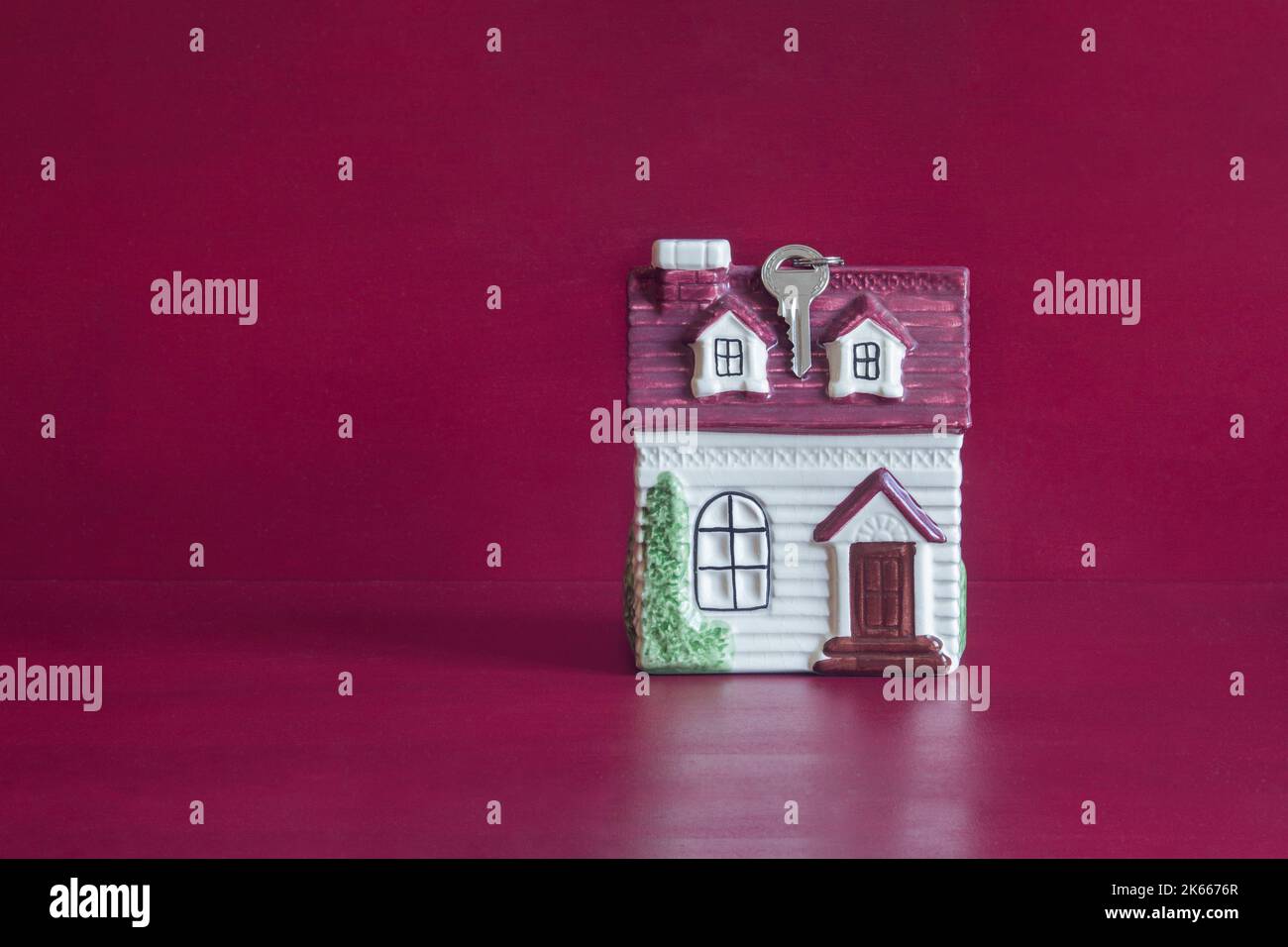 Ein kleines Keramikhaus mit einem Schlüssel, der vom Dach auf einem Hintergrund aus rot lackiertem Holz hängt und einen Kopierraum hat. Real-State-Konzept. Stockfoto