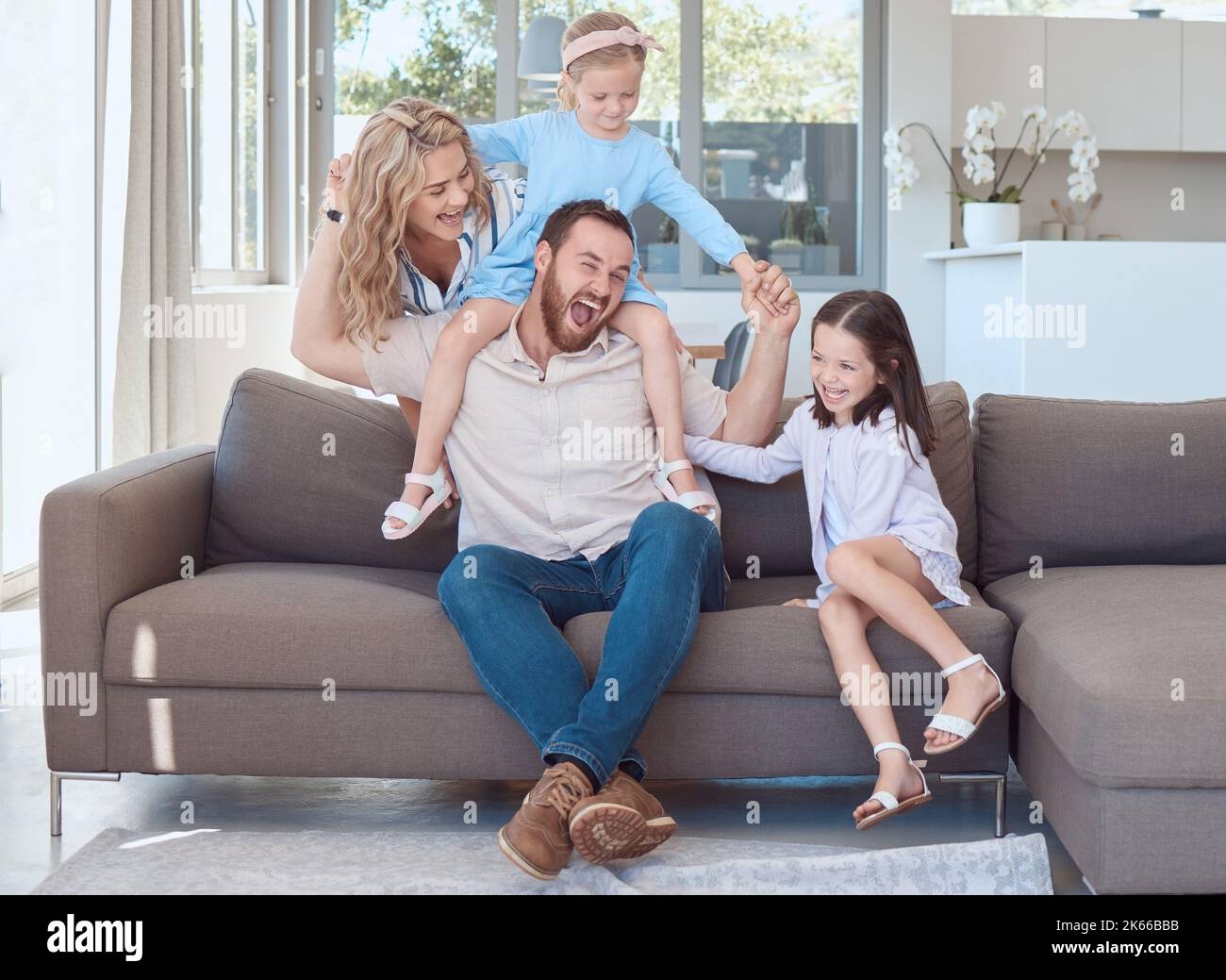 Glückliche junge kaukasische Familie sitzt auf einem Sofa im Wohnzimmer zu Hause und spielt. Entzückende kleine Mädchen und ihre Mutter necken ihren Vater Stockfoto