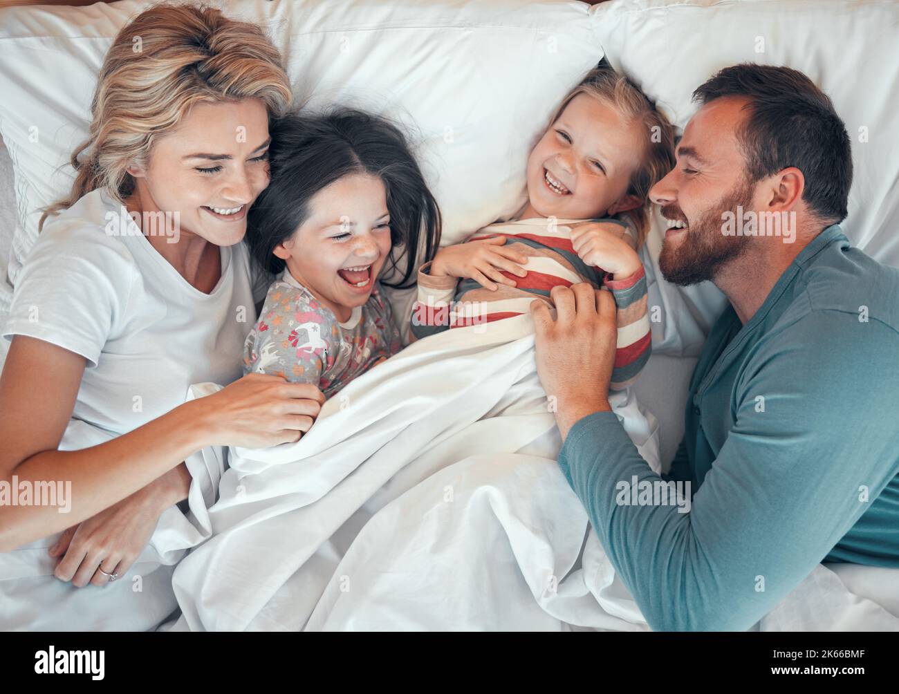 Fröhliche kaukasische Familie, die sich im Bett zusammen entspannt und spielt. Glückliche Eltern kitzeln ihre Kinder, während sie von oben im Bett liegen. Junge Familie mit Stockfoto