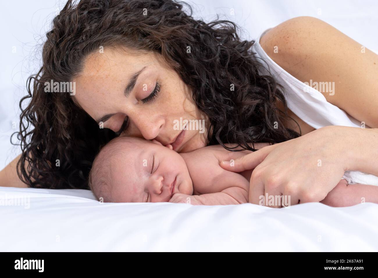 Mutter umarmt und schaut zärtlich auf ihr neugeborenes Baby, während sie schläft und ruht auf dem Bett im Zimmer. Weißer Hintergrund. Familie und Zuneigung Konzept Stockfoto
