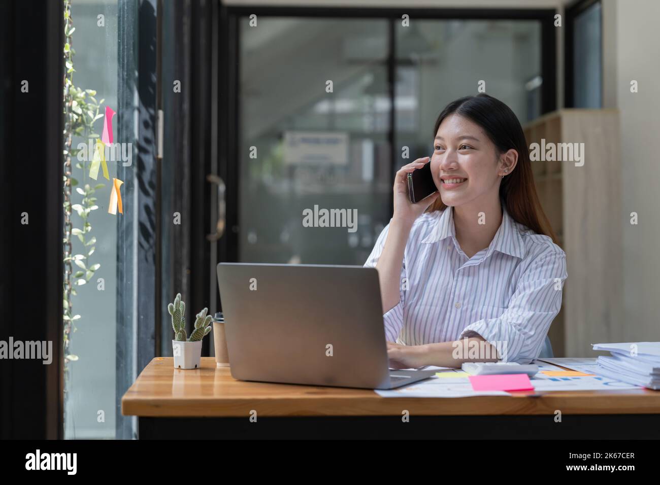 Glückliche junge asiatische Frau, die am Mobiltelefon spricht und lächelt, während sie an ihrem Arbeitsplatz im Büro sitzt. Stockfoto