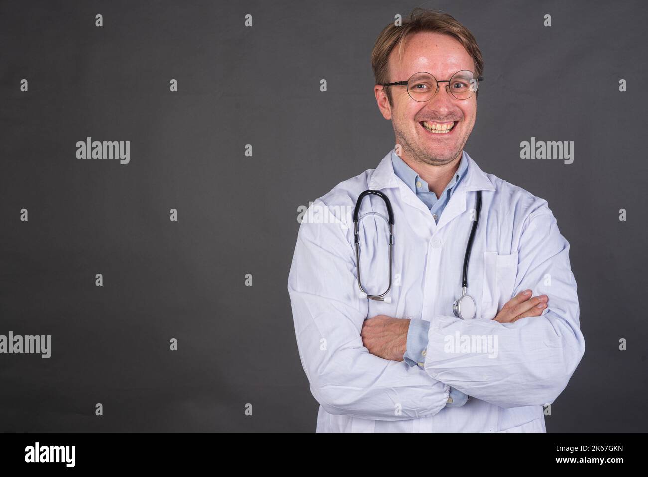 Selbstbewusster männlicher Arzt, der lächelt, mit gekreuzten Armen vor grauem Hintergrund und horizontaler Aufnahme mit Kopierraum Stockfoto