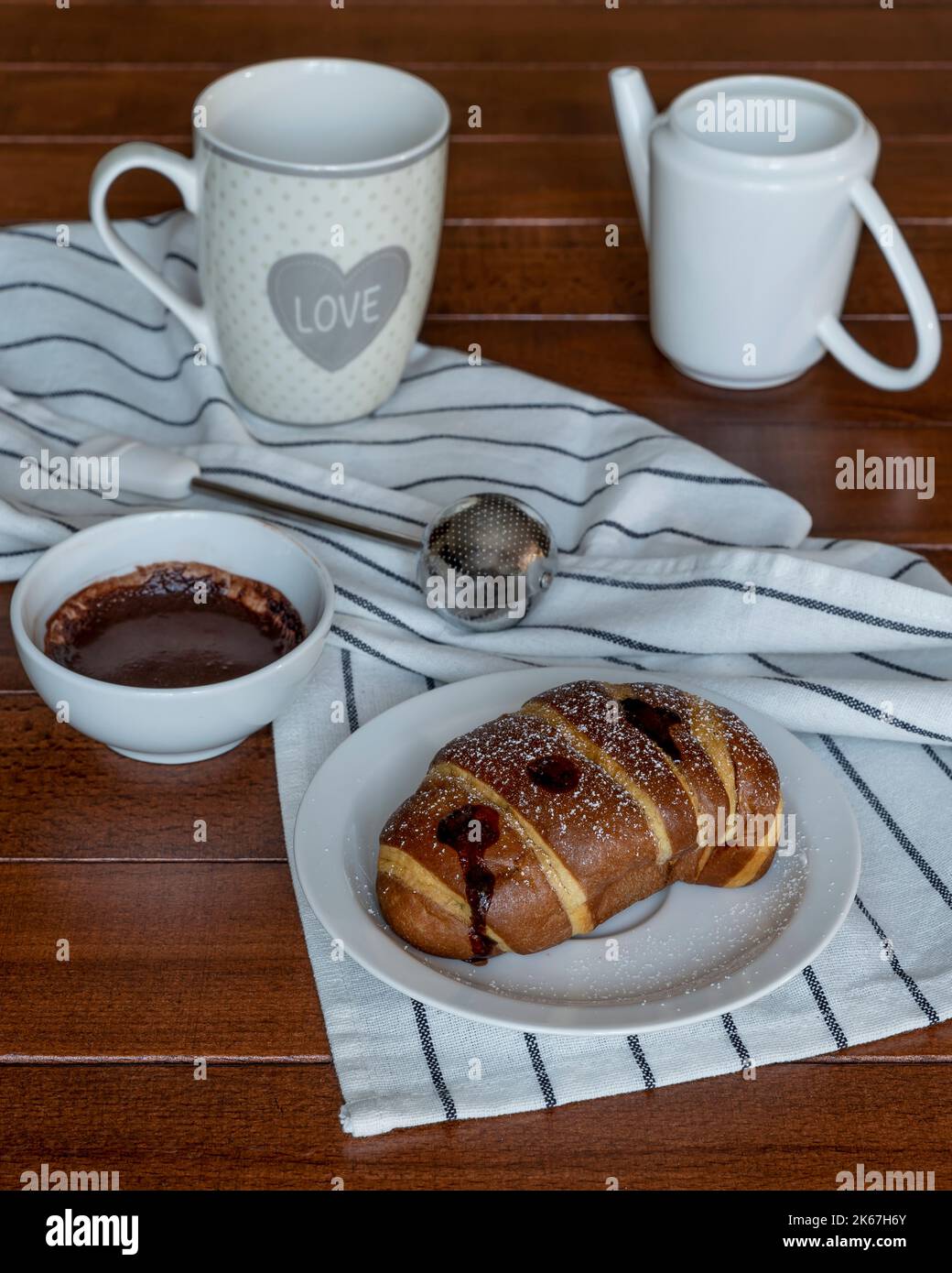 Ein mit Schokolade gefülltes Croissant, bedeckt mit Puderzucker, auf einem Holztisch mit einer weißen Tasse und einem Krug daneben Stockfoto