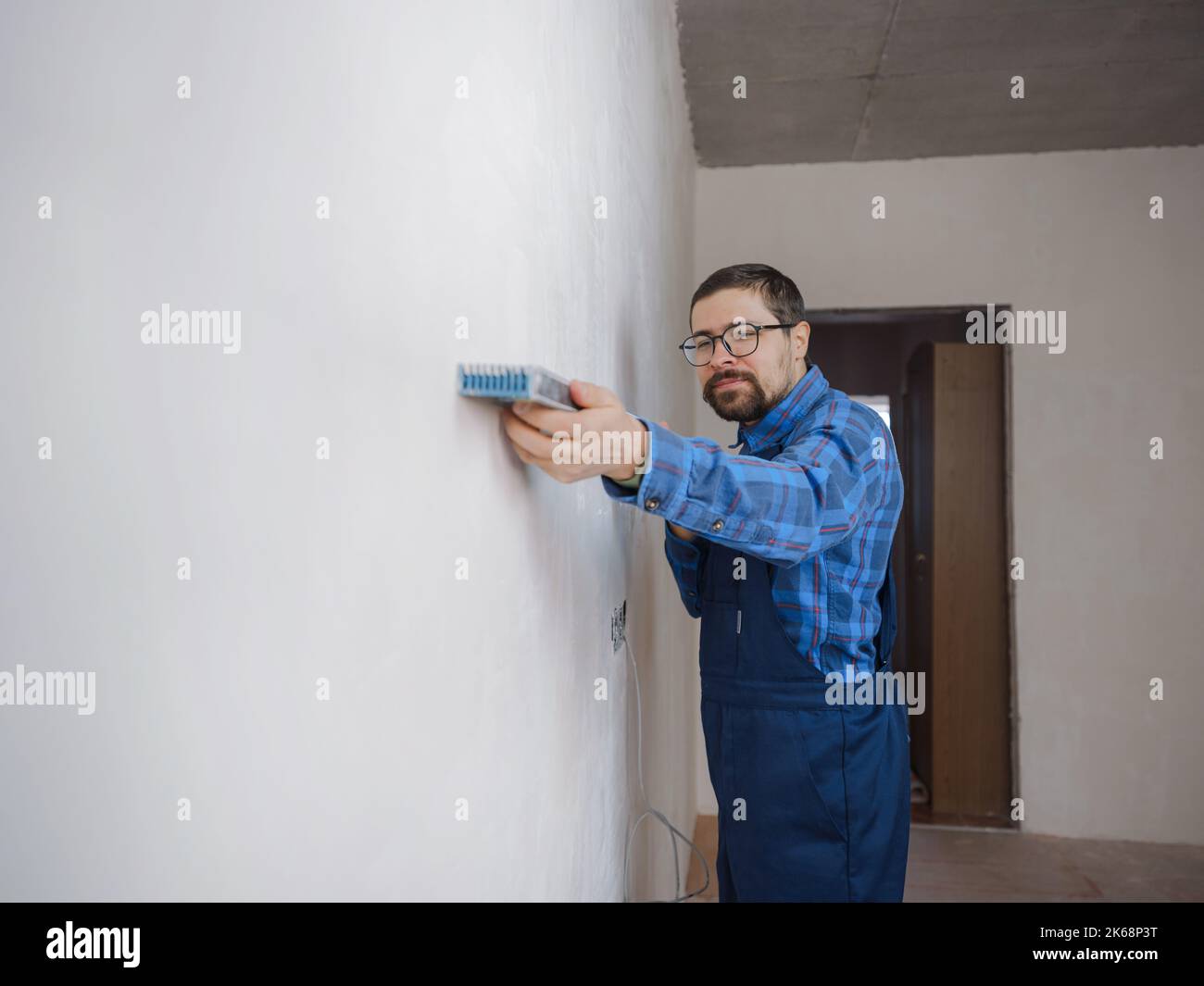 Junger Mann in blau Arbeitsanzug tun Reparatur Wohnung. Renovierungskonzept für das Haus. Kurzer Schuss eines gutaussehenden bärtigen jungen Mannes, der während der Heimverbesserung ein Level-Tool hält Stockfoto