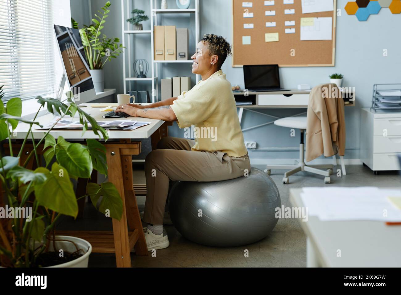 Seitenansicht Porträt einer schwarzen jungen Frau, die am Arbeitsplatz auf dem Fitnessball sitzt und lächelt, sportliche Aktivitäten im Büro Stockfoto