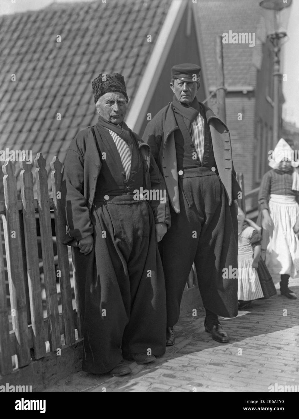 Zwei Männer in traditioneller Kleidung in den Niederlanden c. 1930. Zwei Mädchen stehen hinter ihnen. Männer tragen ein traditionelles Kostüm mit einem langärmeligen Button-up-Hemd zusammen mit einer baggy, plissierten, oft aus Wolle gefertigten Vorderhose, mit silbernen Knöpfen an der quadratischen Frontklappe, Halstuch und Hut – die Form und das Design des Hutes variiert je nach Region. Zu den traditionellen Kostümen gehören in der Regel ein Paar Lederschuhe oder Holzschuhe. Der Stil der traditionellen Kleidung, die in den Niederlanden getragen wurde, begann im 16.. Und 17.. Jahrhundert – ein Vintage-Foto von 1920s/30s. Stockfoto