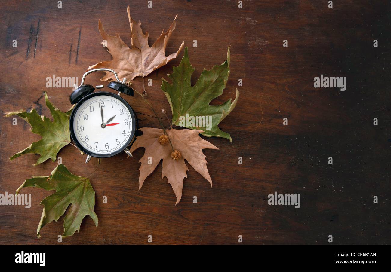 Rückfall, Sommerzeit. Eine Stunde zurück auf gelber Uhr und Herbstlaub auf Holzgrund, Draufsicht Stockfoto