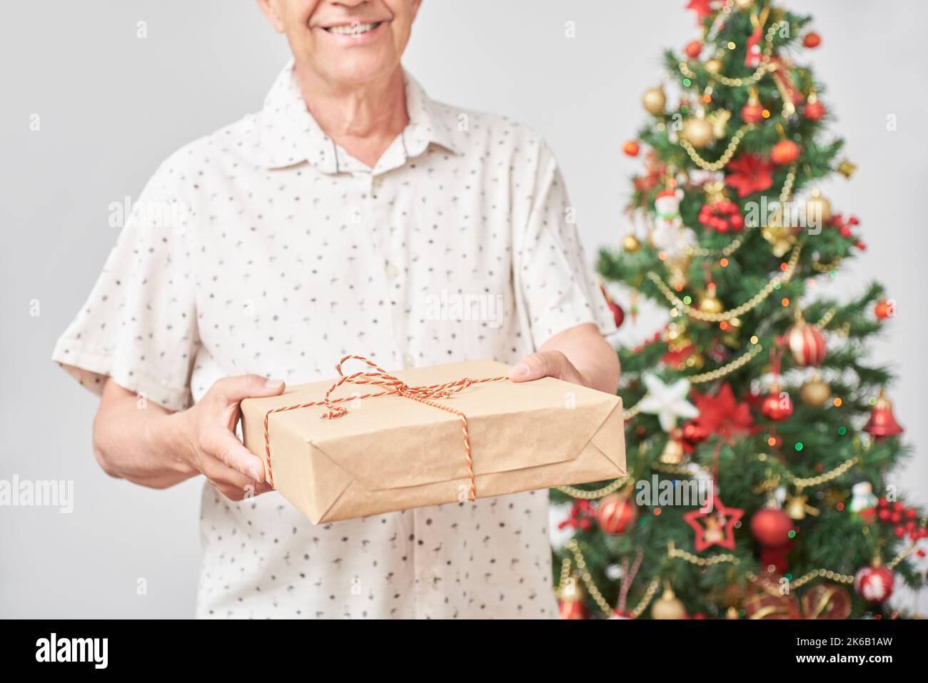 Ein älterer hispanischer Mann lächelt, als er der Kamera ein Weihnachtsgeschenk überreicht. Konzept: Die Freude am Schenken während der Feiertage. Komposition mit Stockfoto