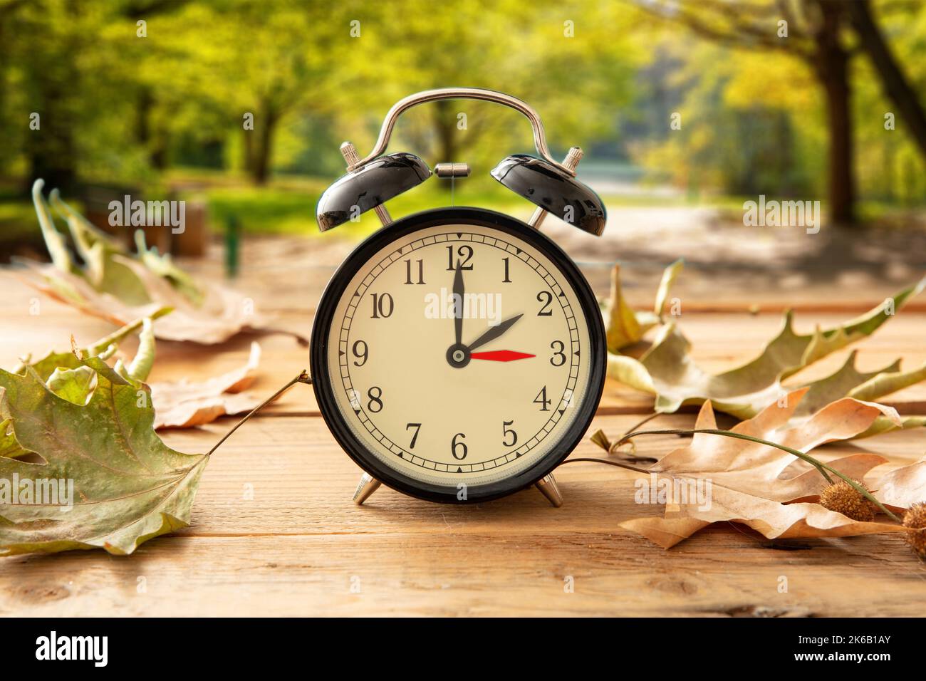 Sommerzeit, Fall eine Stunde zurück. Schwarzer Wecker mit Zeitwechsel auf Holztisch. Herbstbäume und Blätter Hintergrund Stockfoto