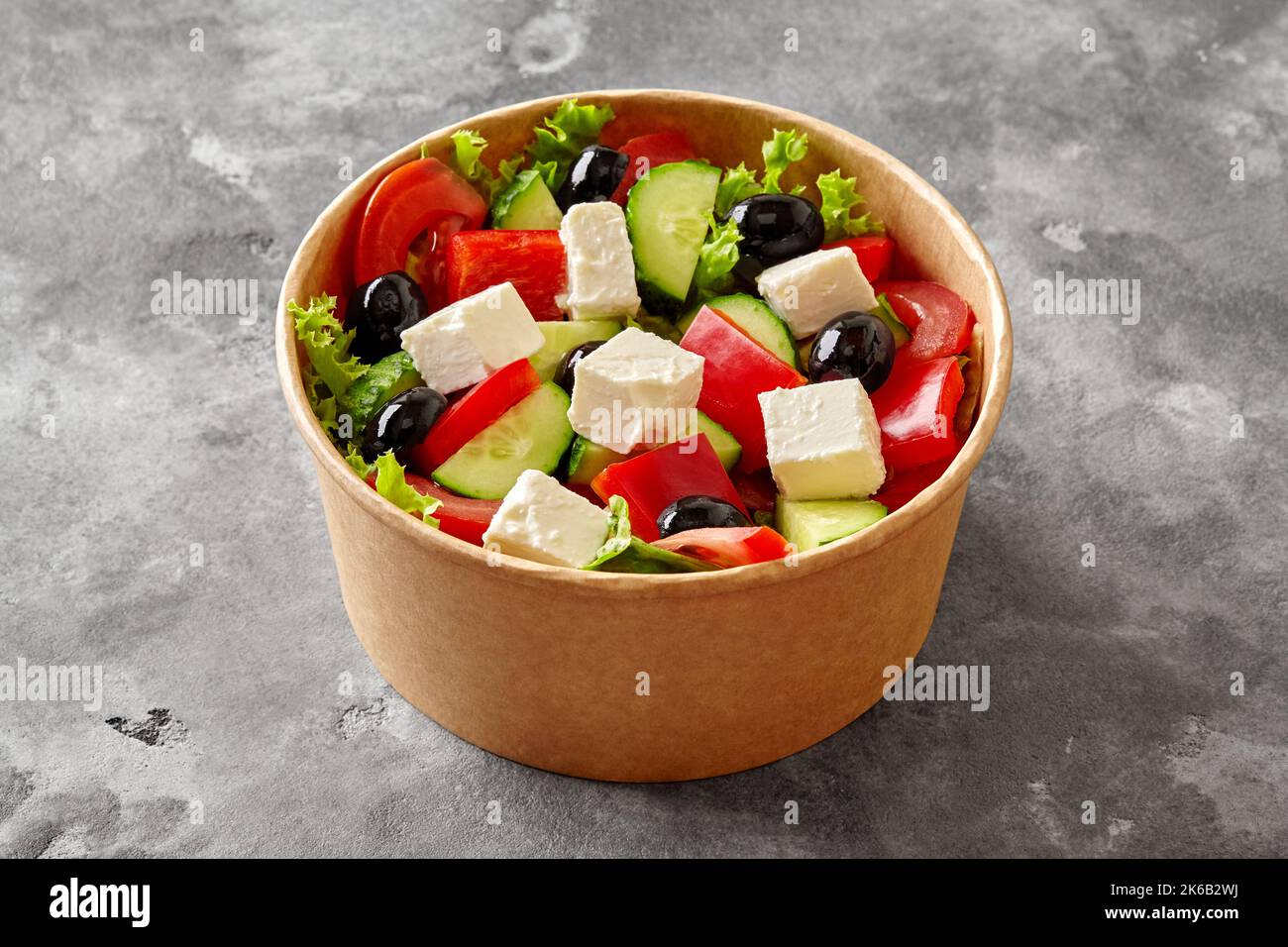 Papierschüssel mit buntem Horiatiki-Salat mit frischem Gemüse, schwarzen Oliven und Feta auf grauer Steinoberfläche Stockfoto