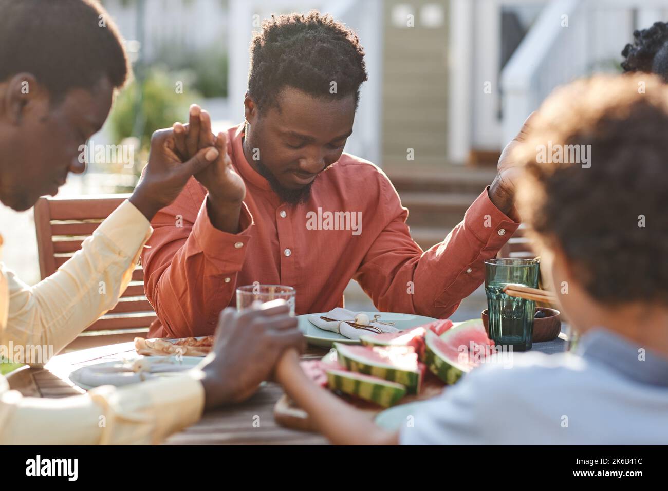 Porträt eines jungen schwarzen Mannes, der im Freien am Tisch Gnade sagt, während die Familie zusammenkommt und die Hände im Sonnenlicht hält Stockfoto